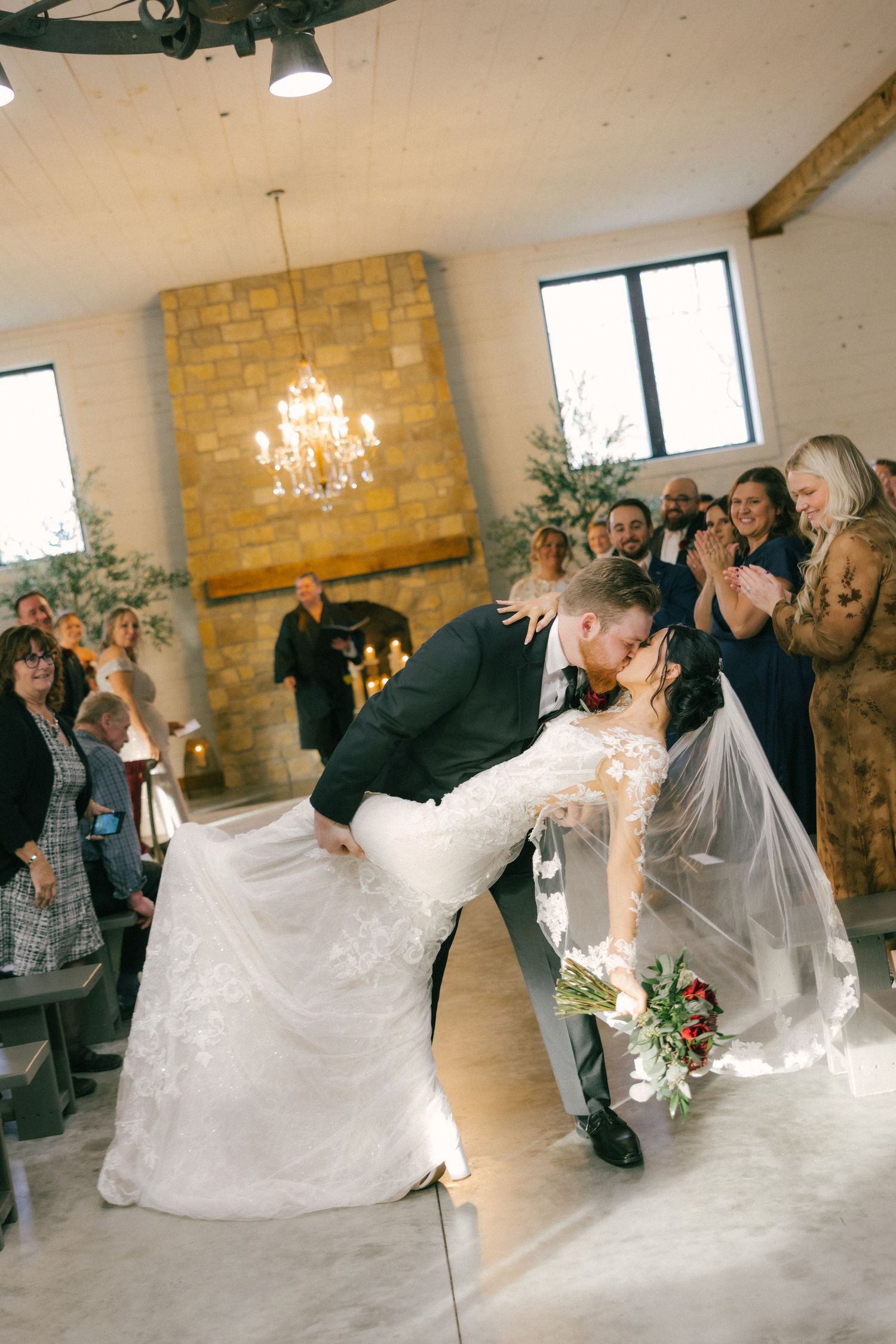 A wedding couple performs a dramatic dip dance move in an elegant reception venue with exposed brick walls.