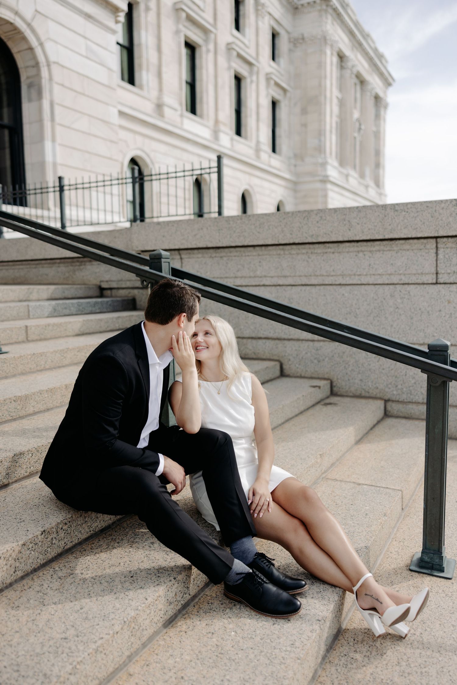 Romantic couple in formal attire sitting together on concrete steps of an elegant white stone building.
