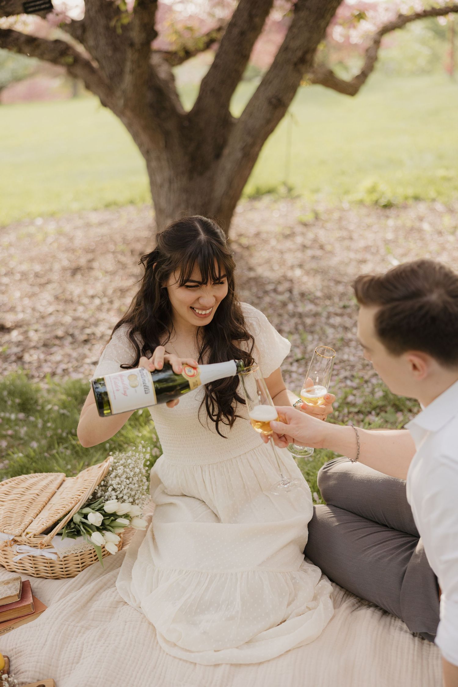 Couple enjoying a romantic spring picnic under a blooming tree with wine and a picnic basket.