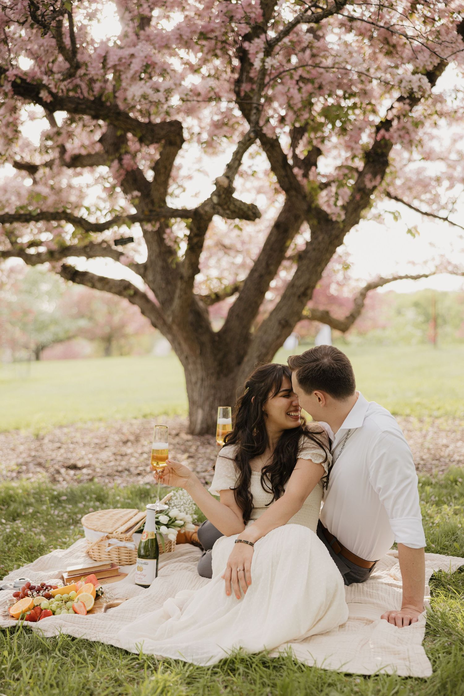 A couple enjoys a romantic spring picnic under blooming cherry blossoms with wine and snacks on a white blanket.