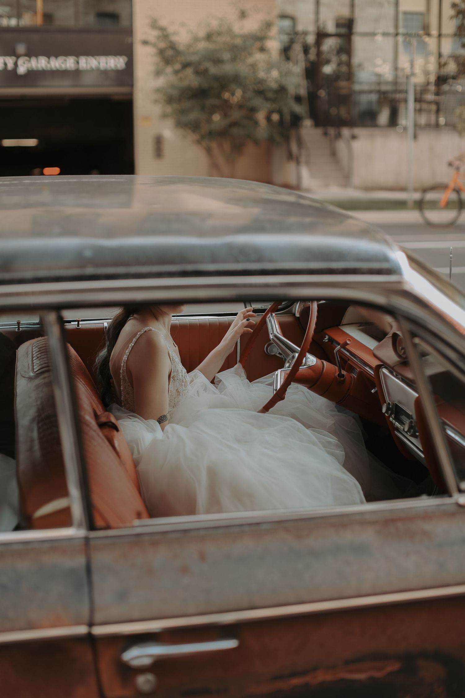 A bride in a white wedding dress sits in the driver's seat of a vintage car with brown leather interior.