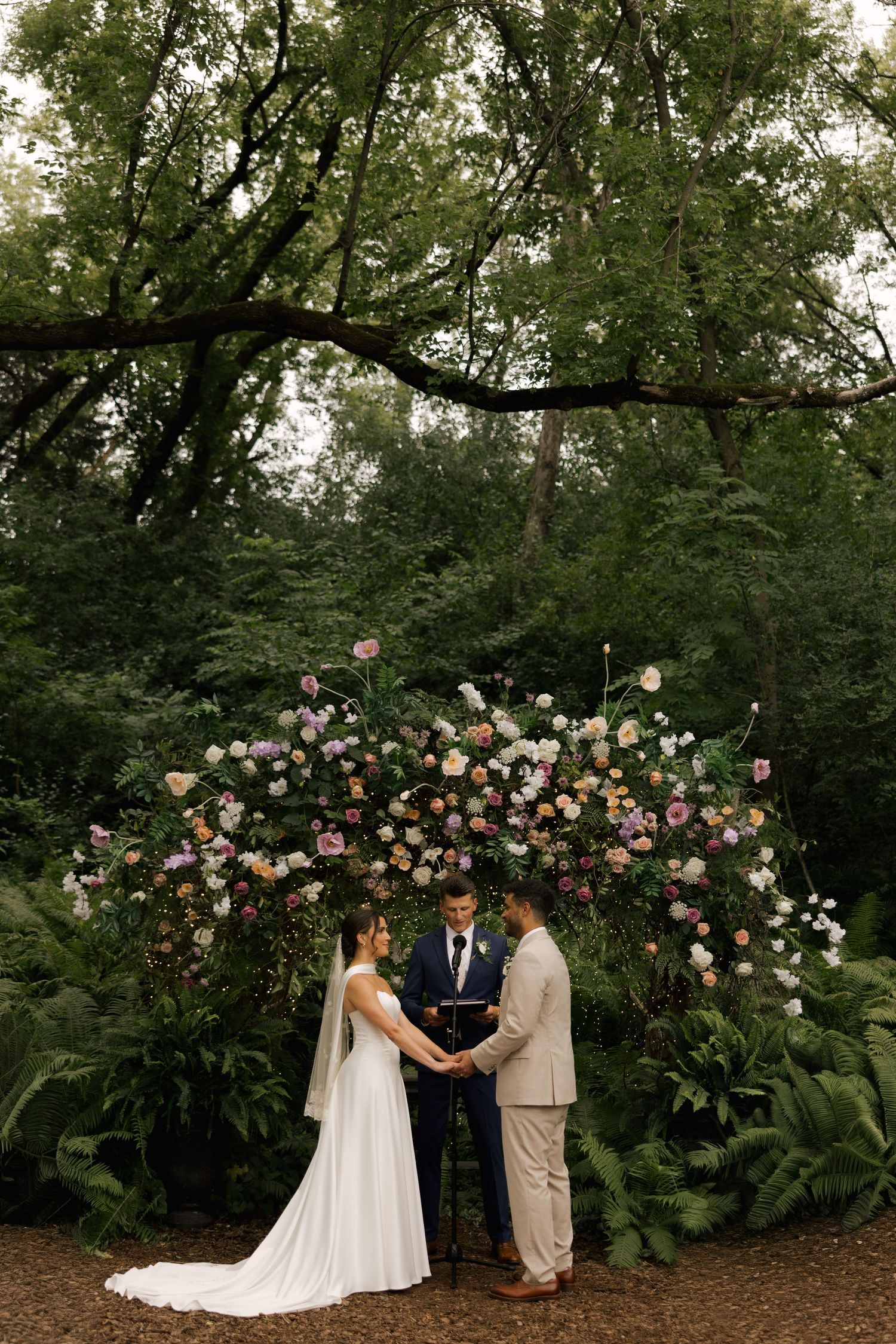 Couple exchanges vows under floral arch surrounded by lush greenery in an outdoor forest wedding ceremony.