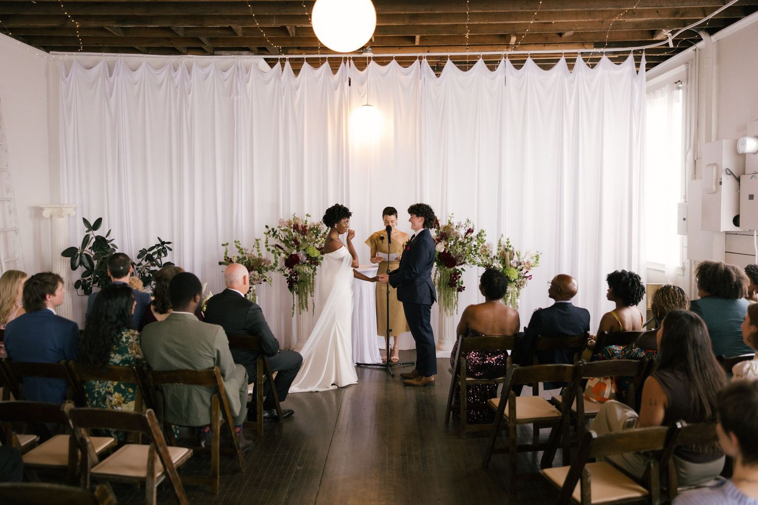 Couple exchanges vows at an intimate indoor wedding ceremony with floral arrangements and seated guests in a modern venue.