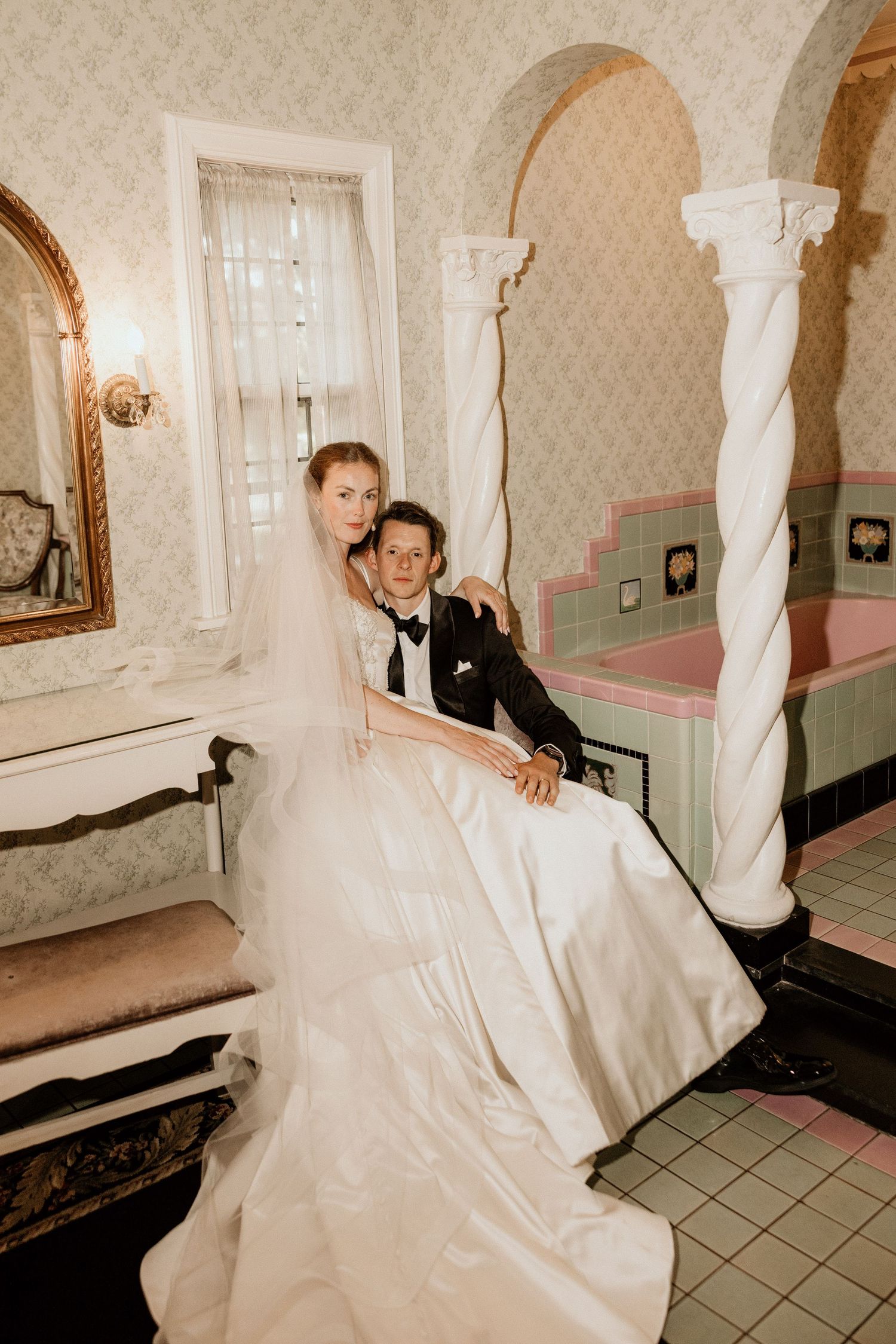 Bride in white gown and veil sits with groom in tuxedo in elegant vintage bathroom with pink tub.