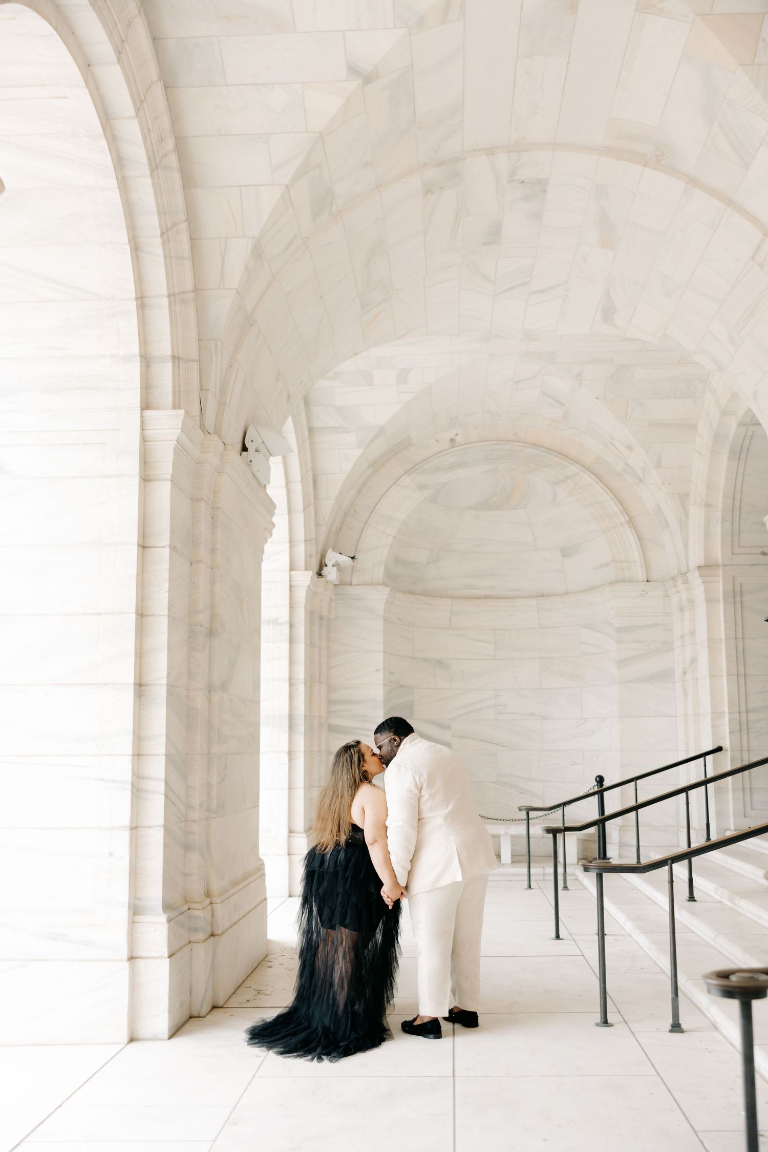 A person in a black dress sits on white marble stairs in an elegant architectural space with arched columns.