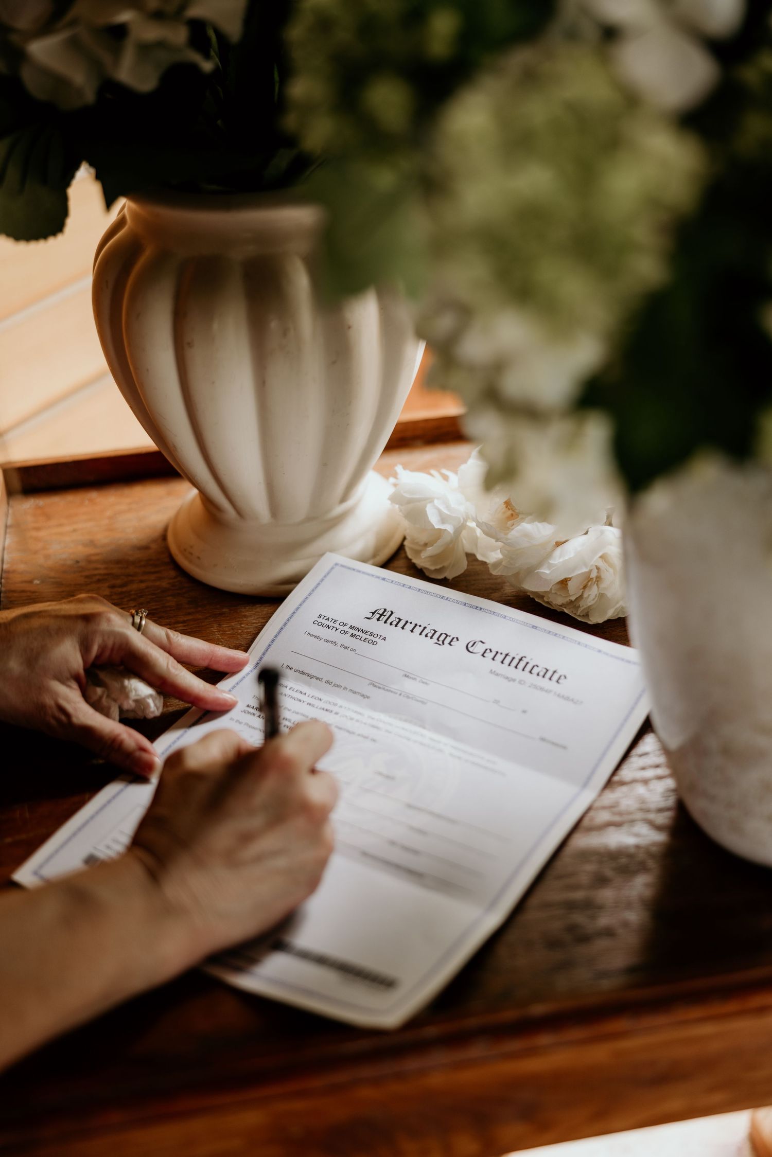 A hand signs a marriage certificate beside a white vase and flowers on a wooden table in warm lighting.