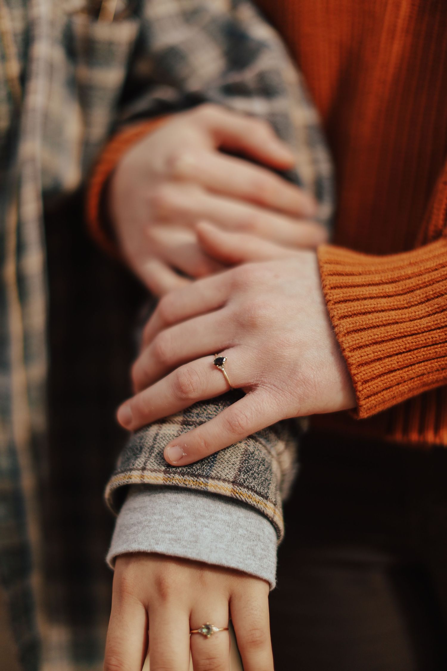 Close-up of hands wearing engagement rings with orange sweater and plaid fabric.