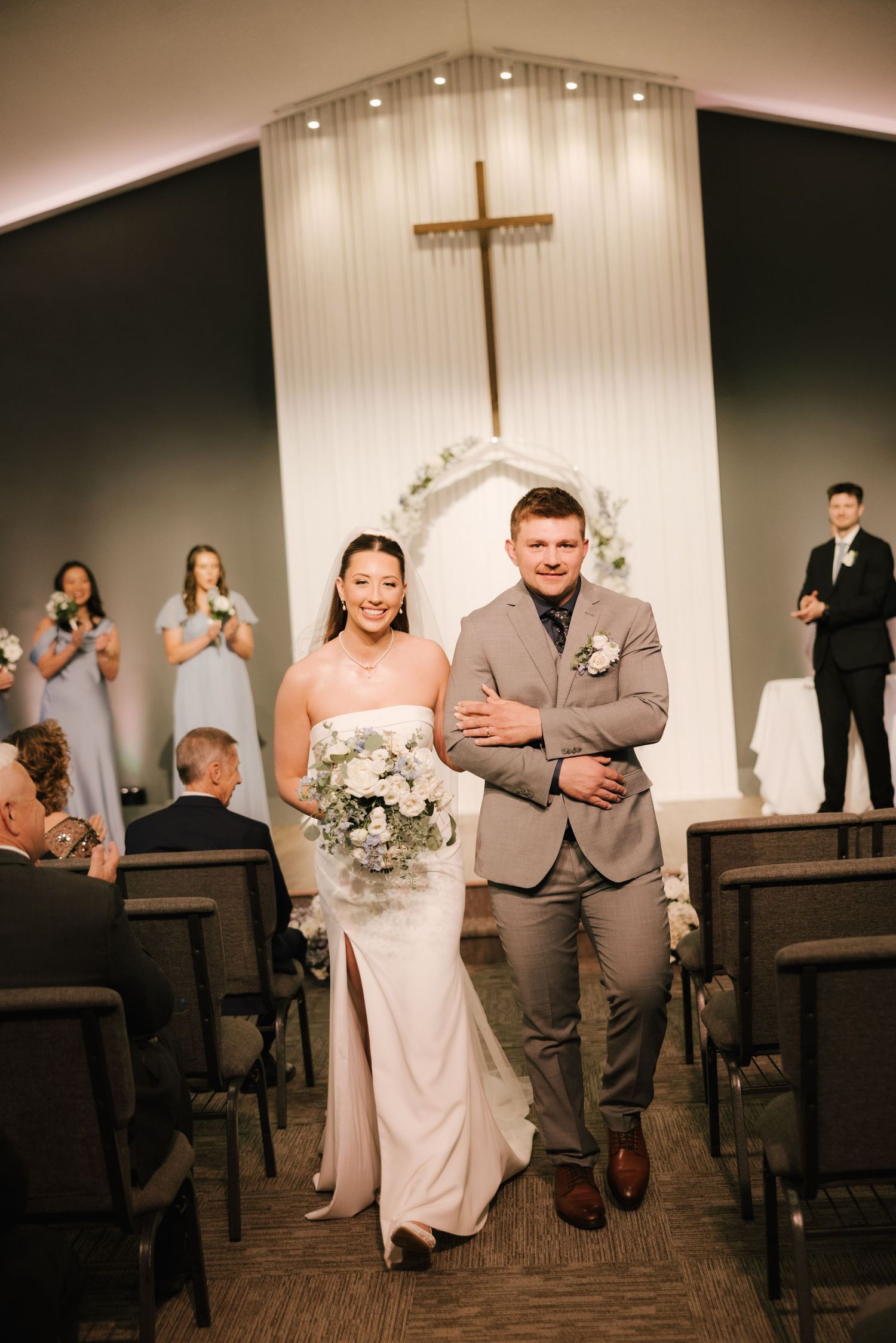 Bride and groom walk down the aisle together in a church wedding ceremony, with a cross and floral arch in the background.