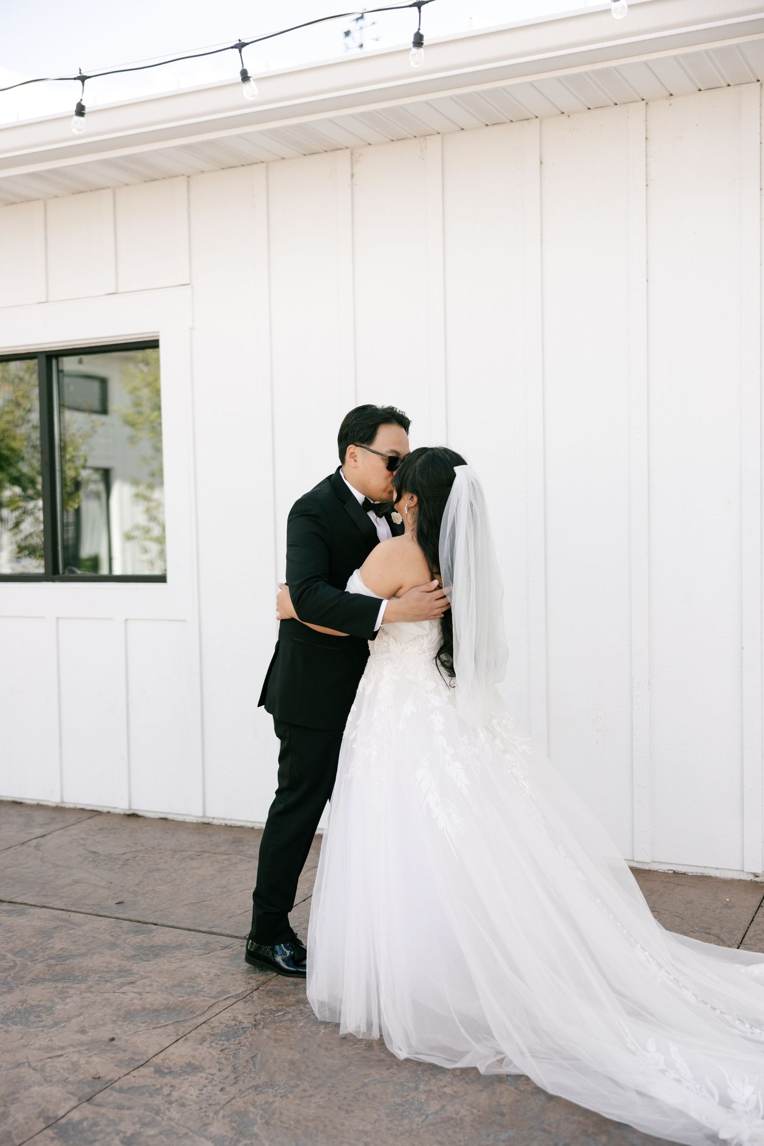 A bride and groom share a romantic kiss outside a white barn venue, with the bride in a flowing white gown and veil.