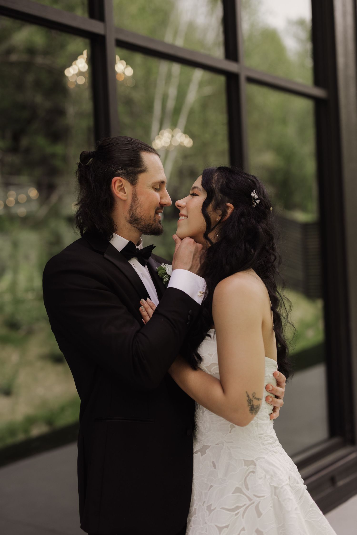 A bride and groom share a romantic moment, nearly kissing, dressed in a white gown and black tuxedo before large windows.