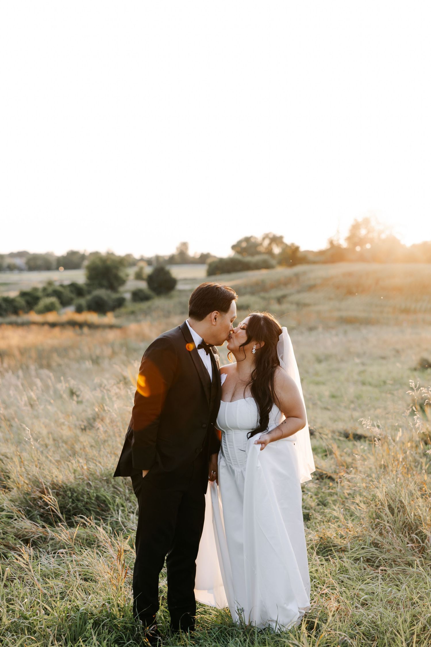 A couple in wedding attire share an intimate moment in a sunlit meadow at golden hour.