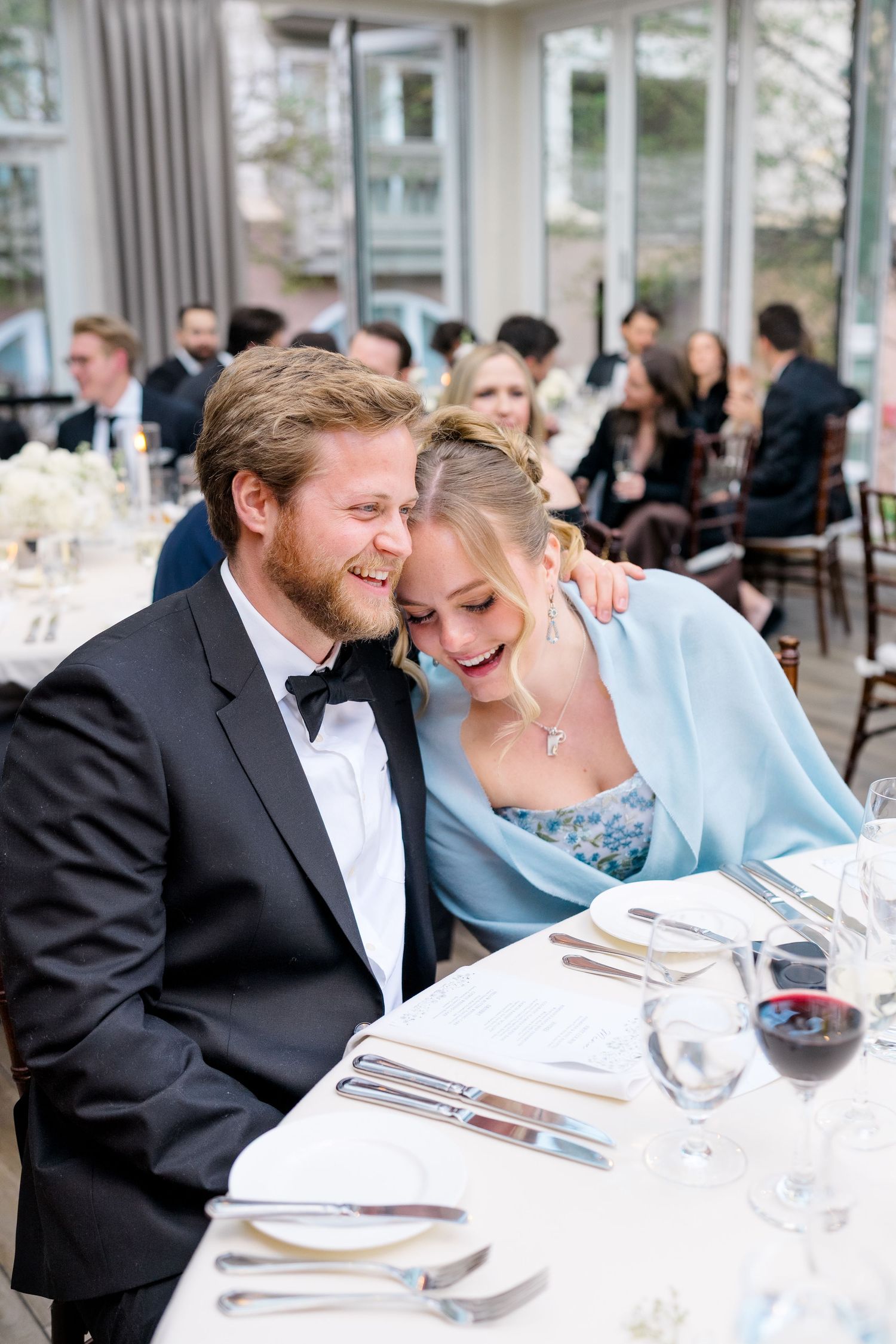 Couple laughing together at elegant wedding reception dinner table, man in tuxedo, woman in light blue dress.