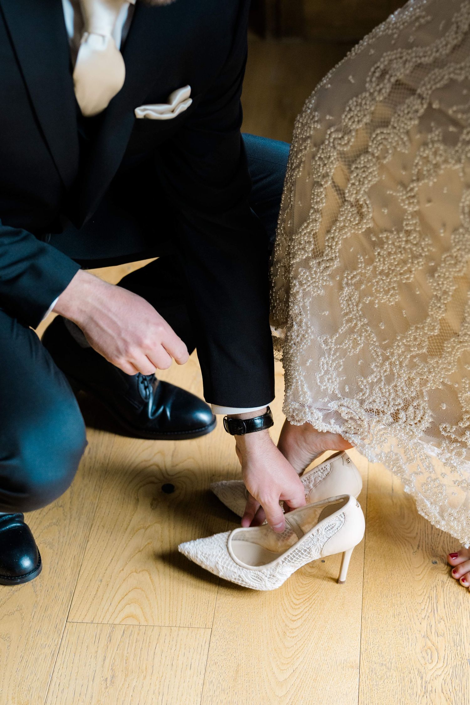 Groom in black suit kneeling to help bride put on white lace heels on wooden floor during wedding preparation.