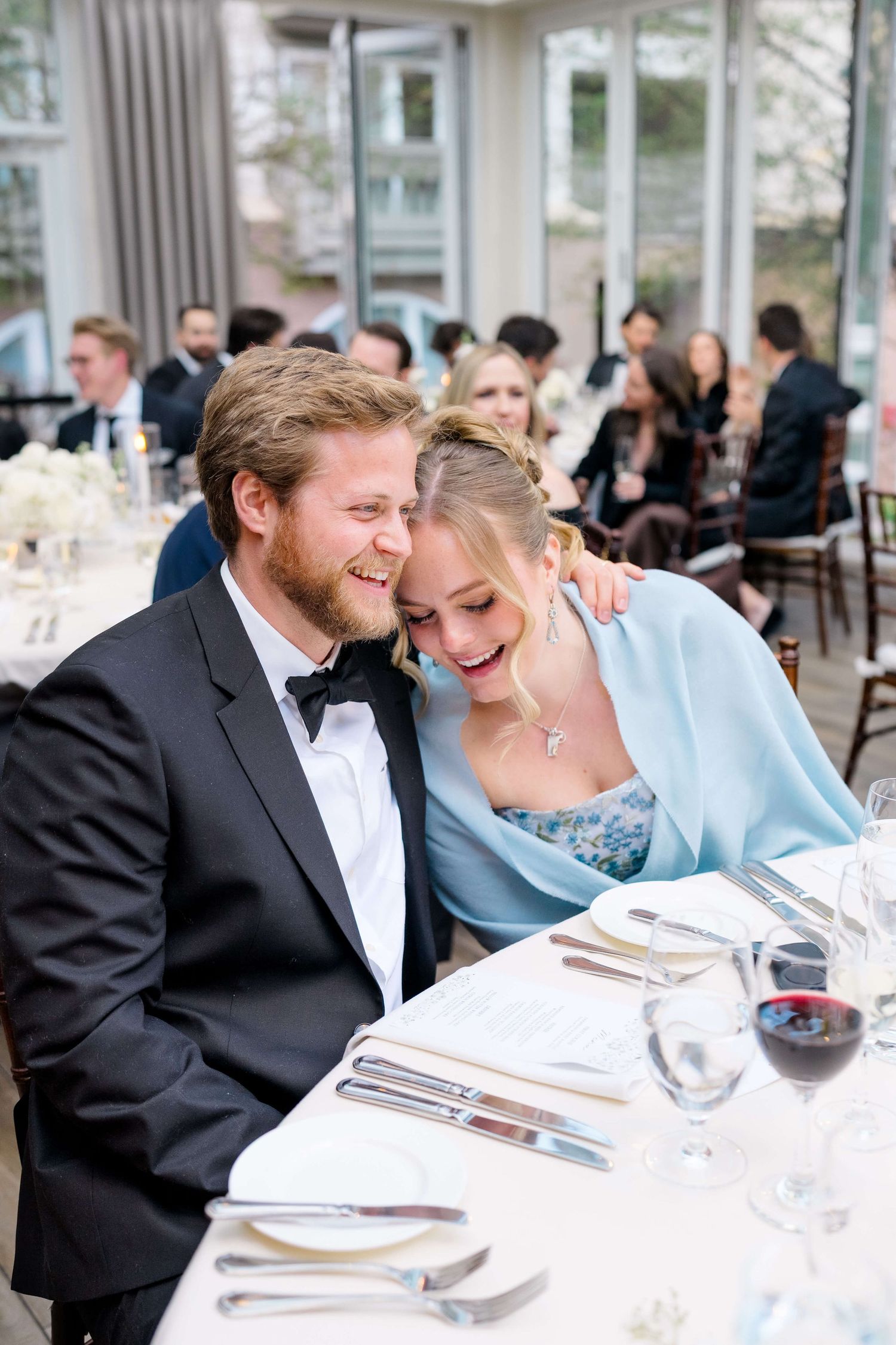 Couple laughing together at elegant wedding reception dinner table with wine glasses and white floral centerpieces.