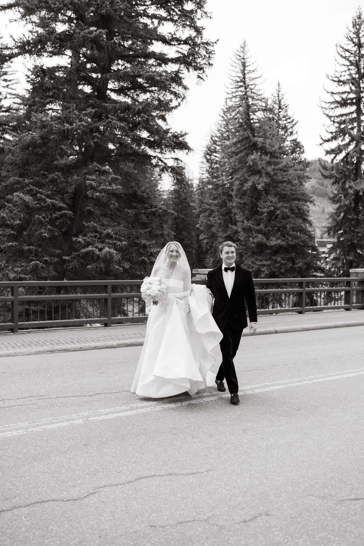 Black and white photo of bride and groom walking across a road, surrounded by tall pine trees on their wedding day.