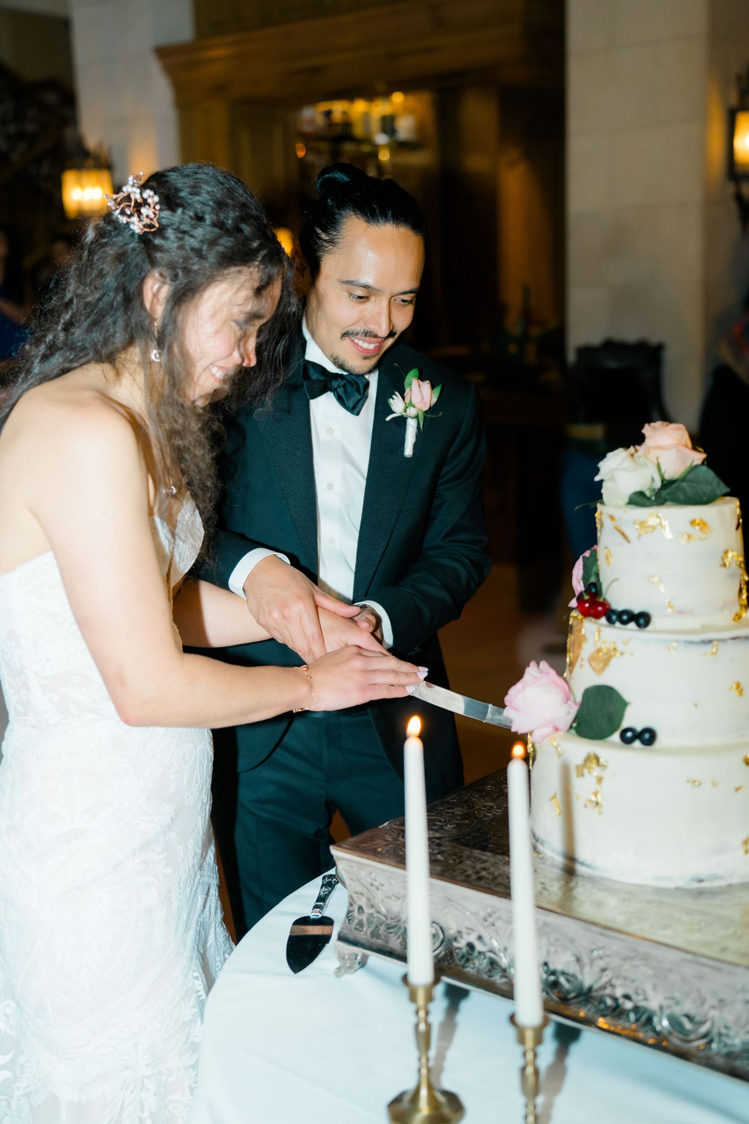 A bride and groom cut their elegant white wedding cake together at their reception, with candlesticks on the table.