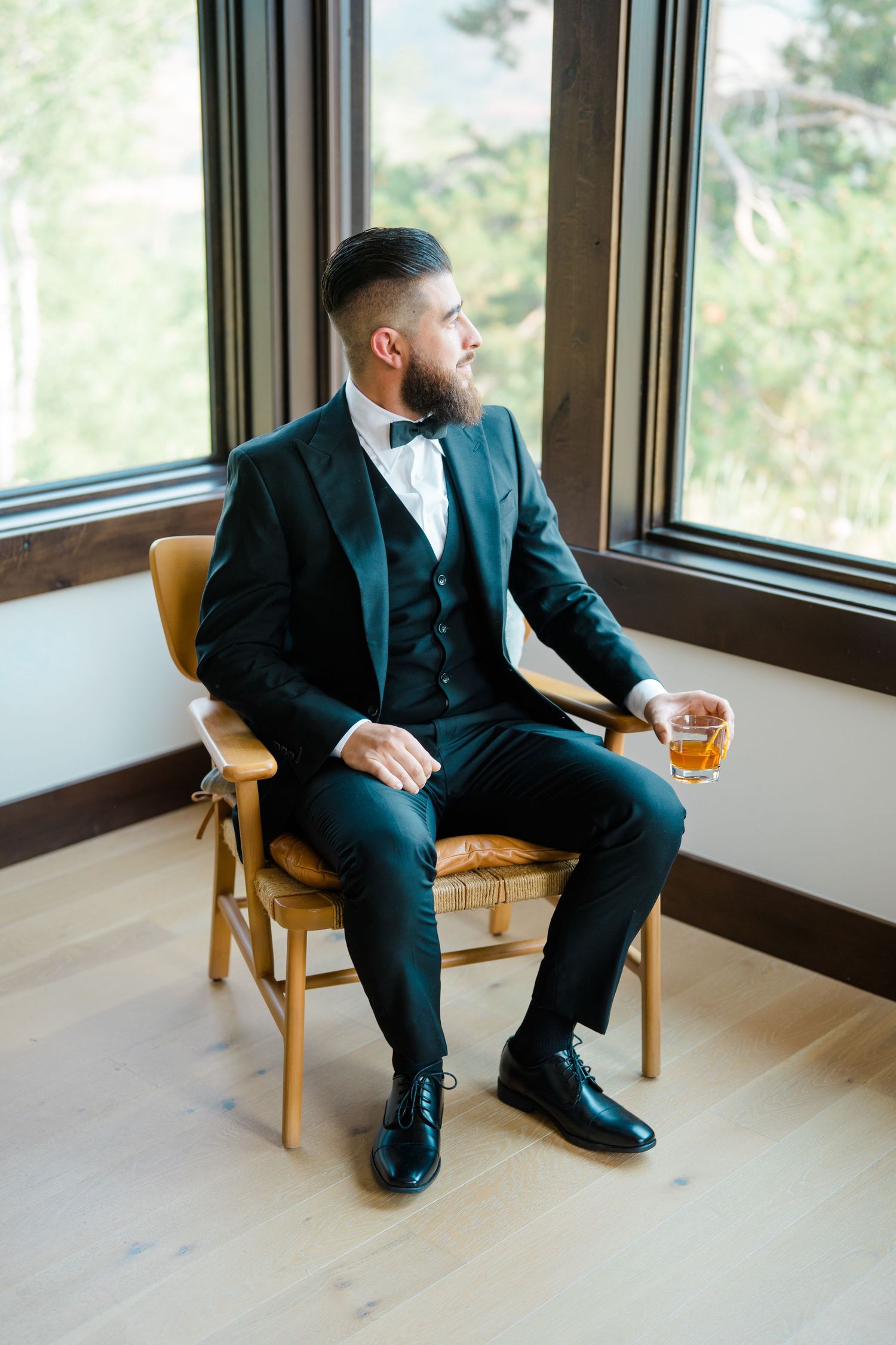 A groom in a dark suit sits contemplatively in a wooden chair by large windows.