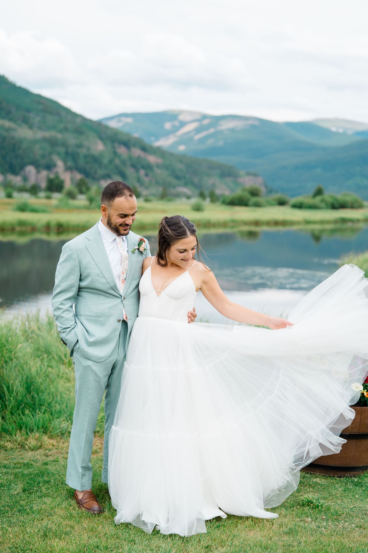 A couple poses together in a scenic mountain landscape with a lake, the bride's white dress flowing in the breeze.