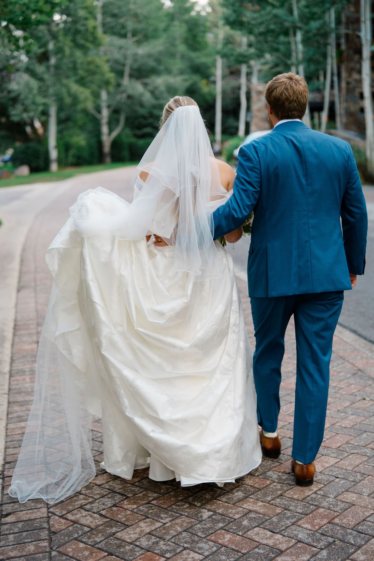 Wedding couple walking on brick path through garden with flowing white dress and navy suit.