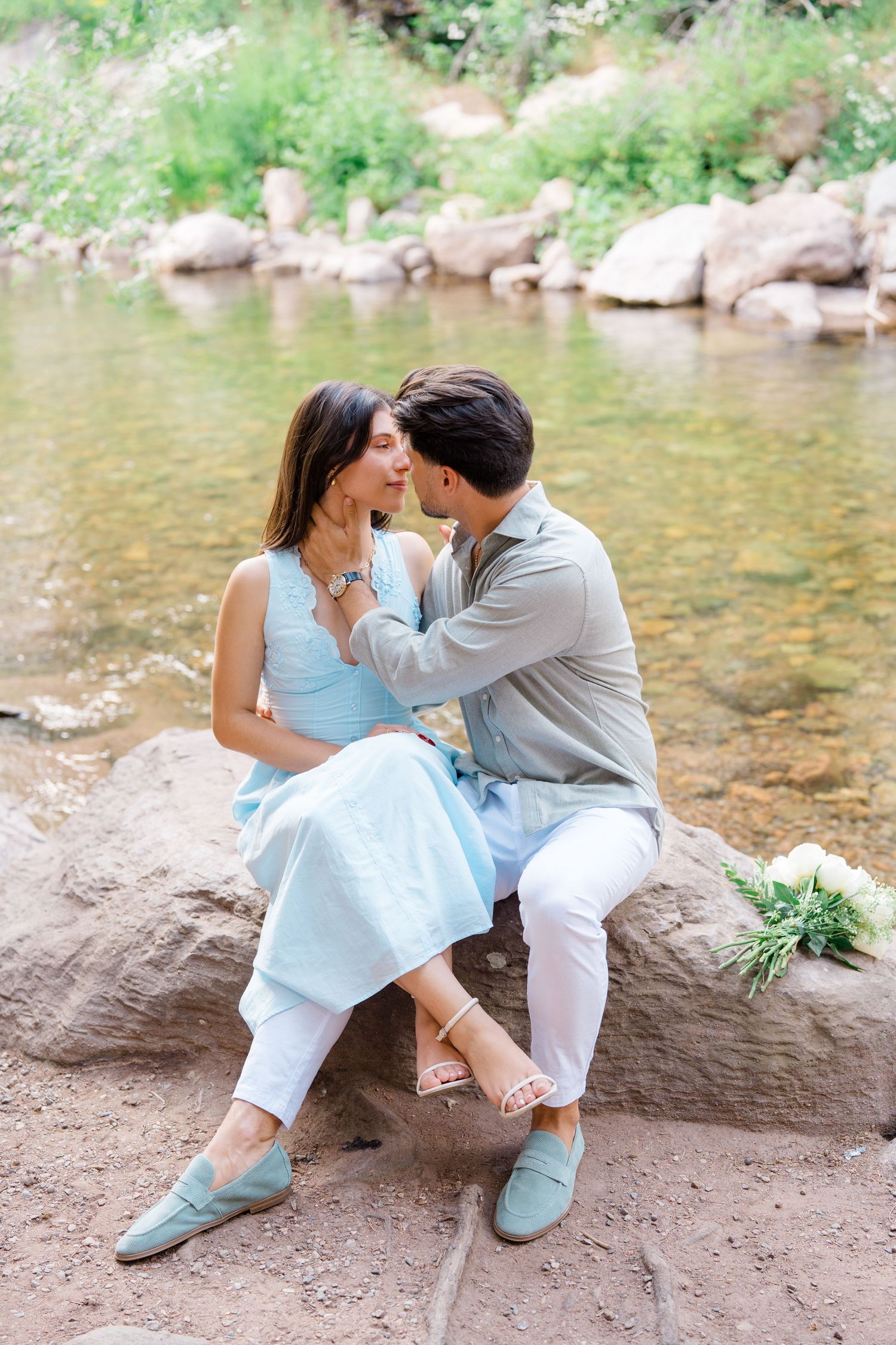 A couple in light blue and white outfits sits together by a scenic stream with rocks and greenery.