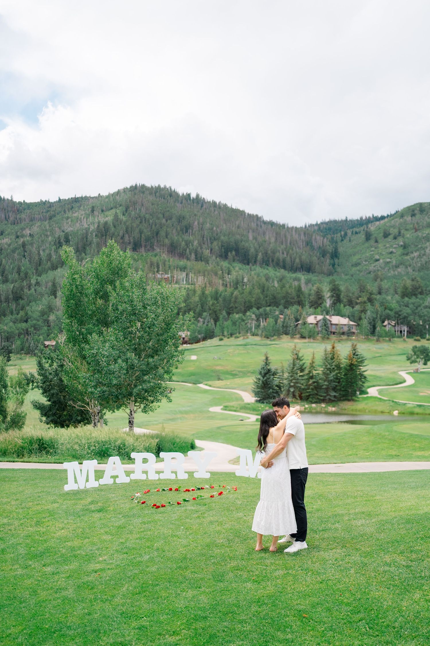 Scenic mountain wedding ceremony setup with white chairs on a lush green golf course surrounded by evergreen trees.