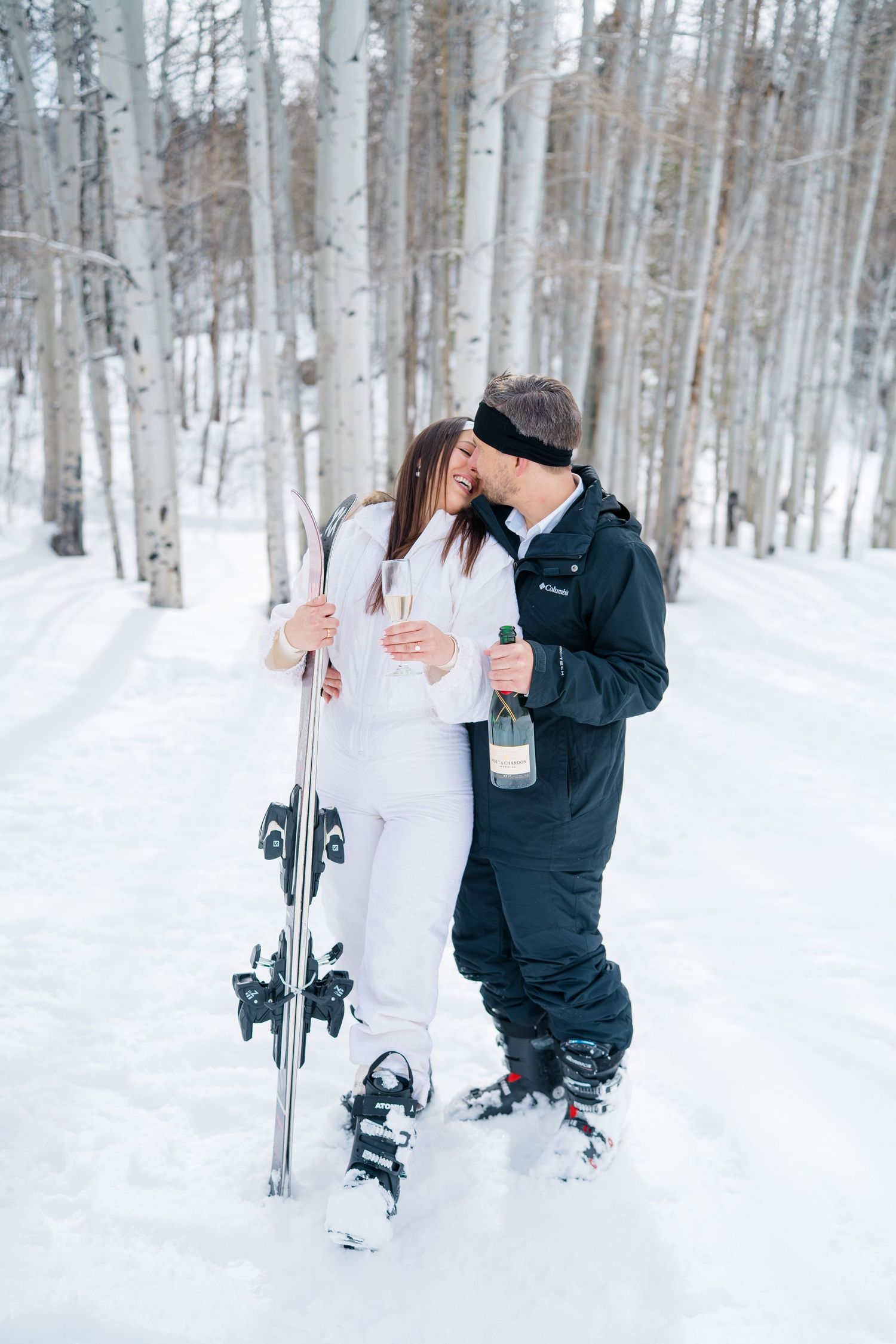 Winter wedding couple stands in snowy aspen forest holding skis and sharing a romantic moment together.