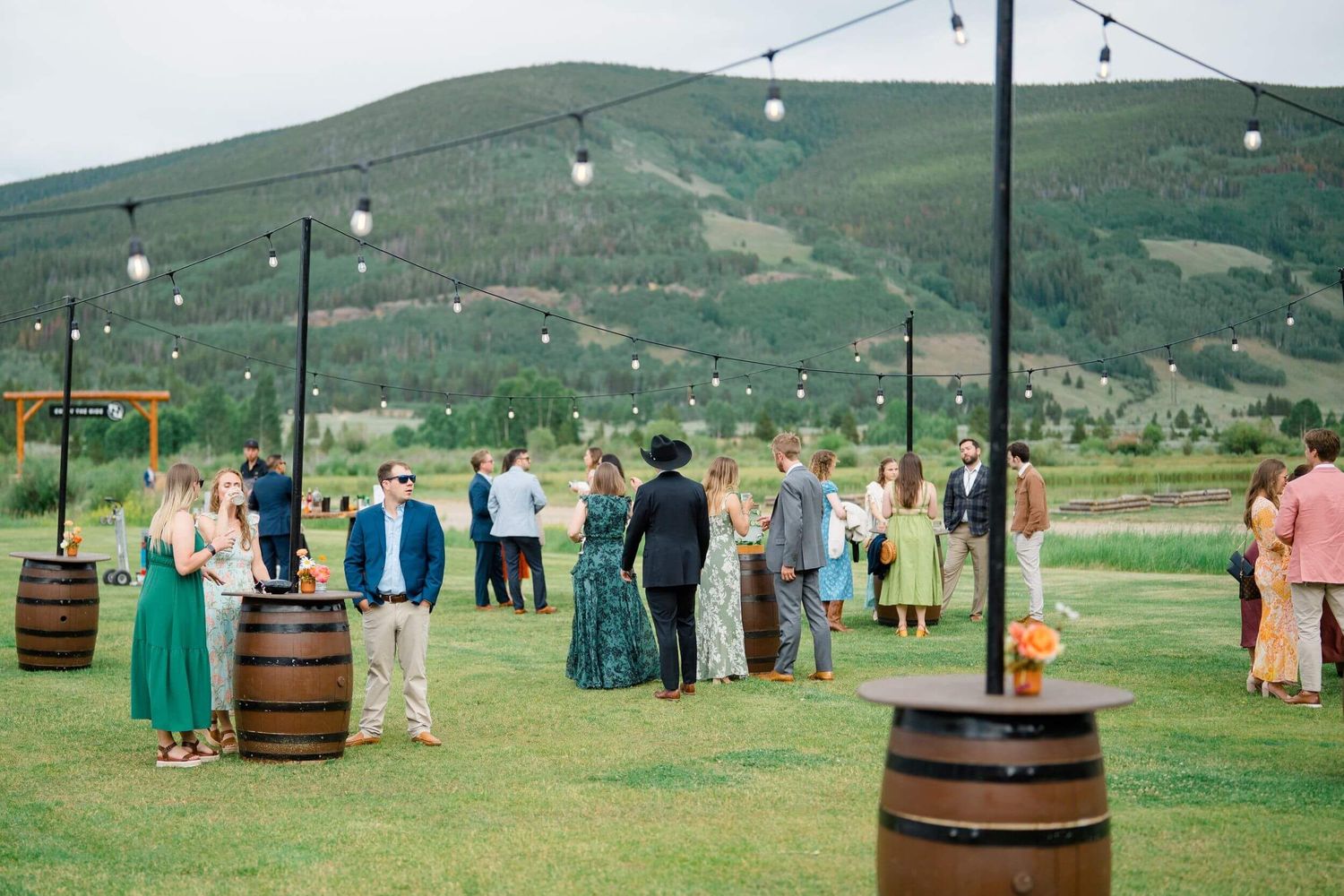 Camp Hale Outdoor wedding cocktail hour with guests mingling around wine barrel tables under string lights against mountain backdrop.