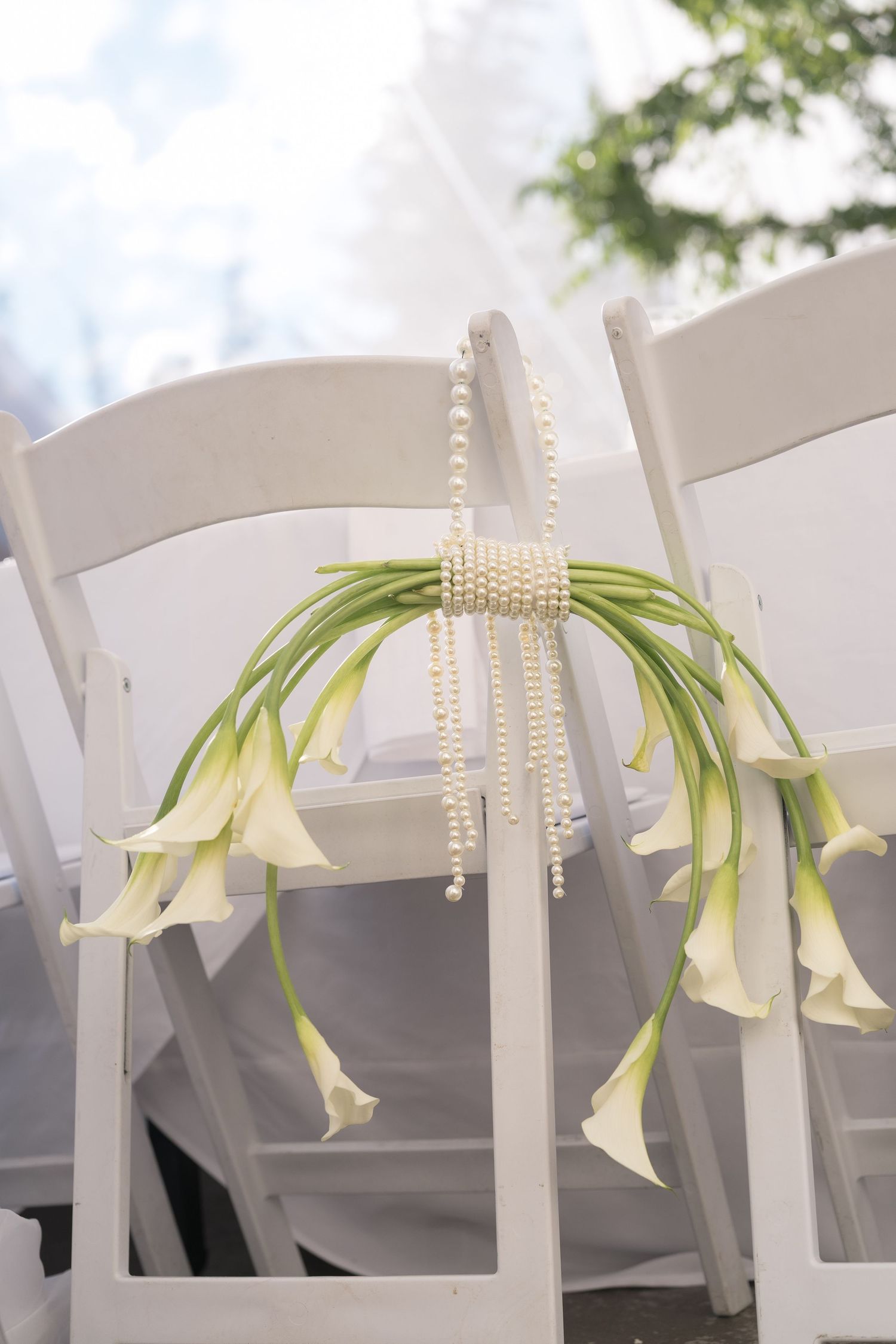 White calla lilies adorned with pearl strands decorating a white folding chair at an outdoor wedding ceremony.