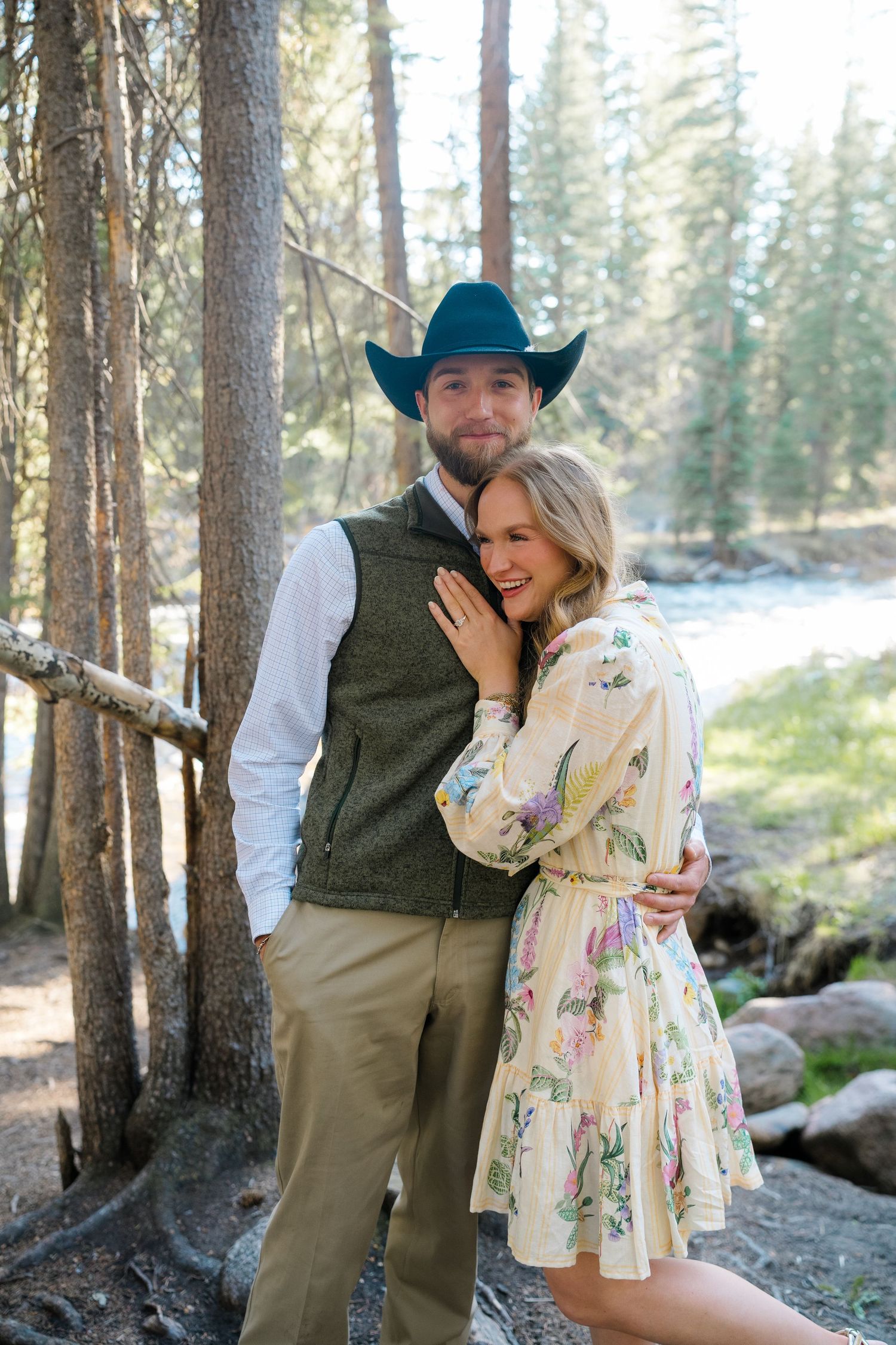 Couple posing in forest, man in cowboy hat and vest, woman in floral dress, embracing near a river.
