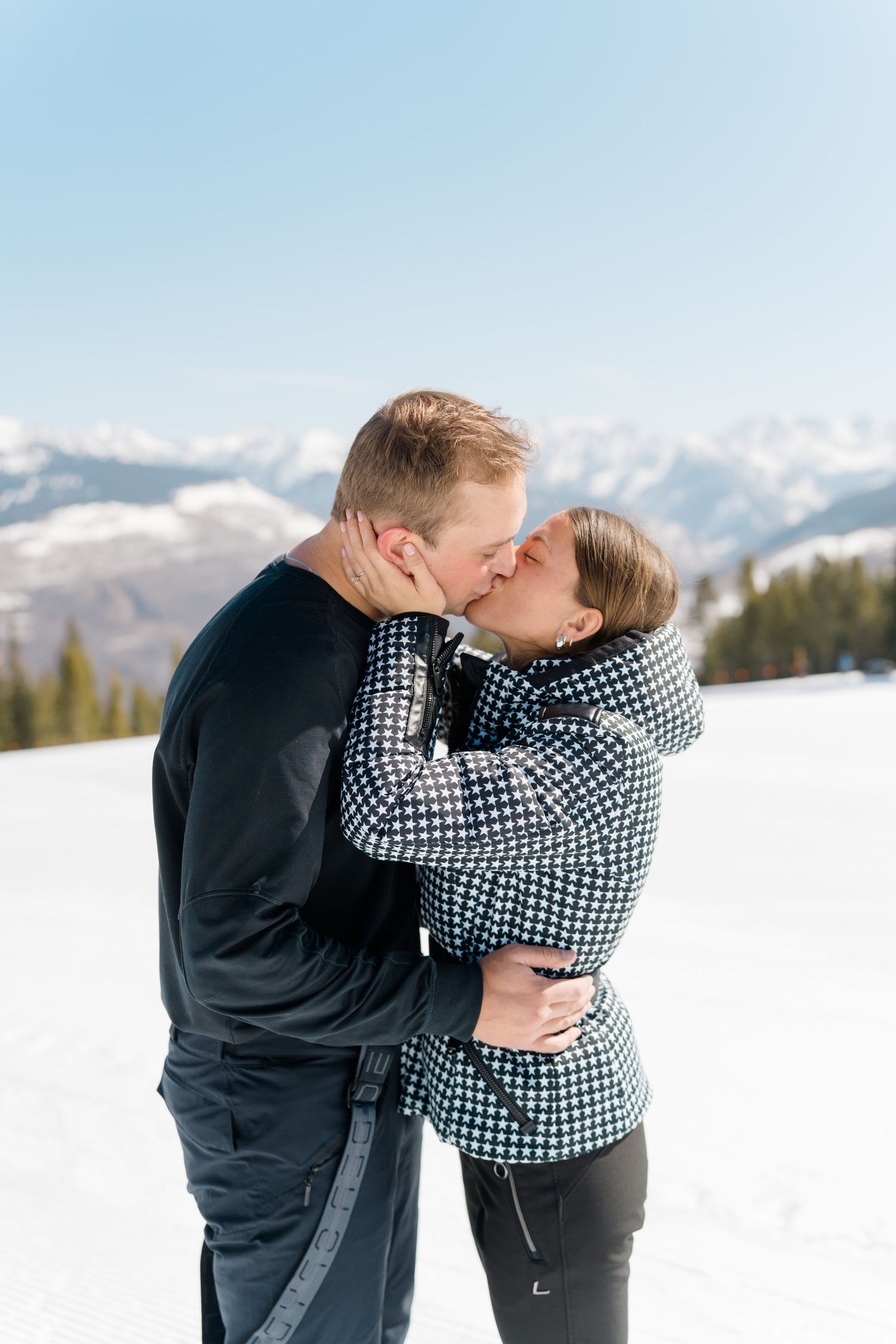 A couple shares a romantic kiss in the snow with snow-capped mountains visible in the background during vail proposal engagement session.