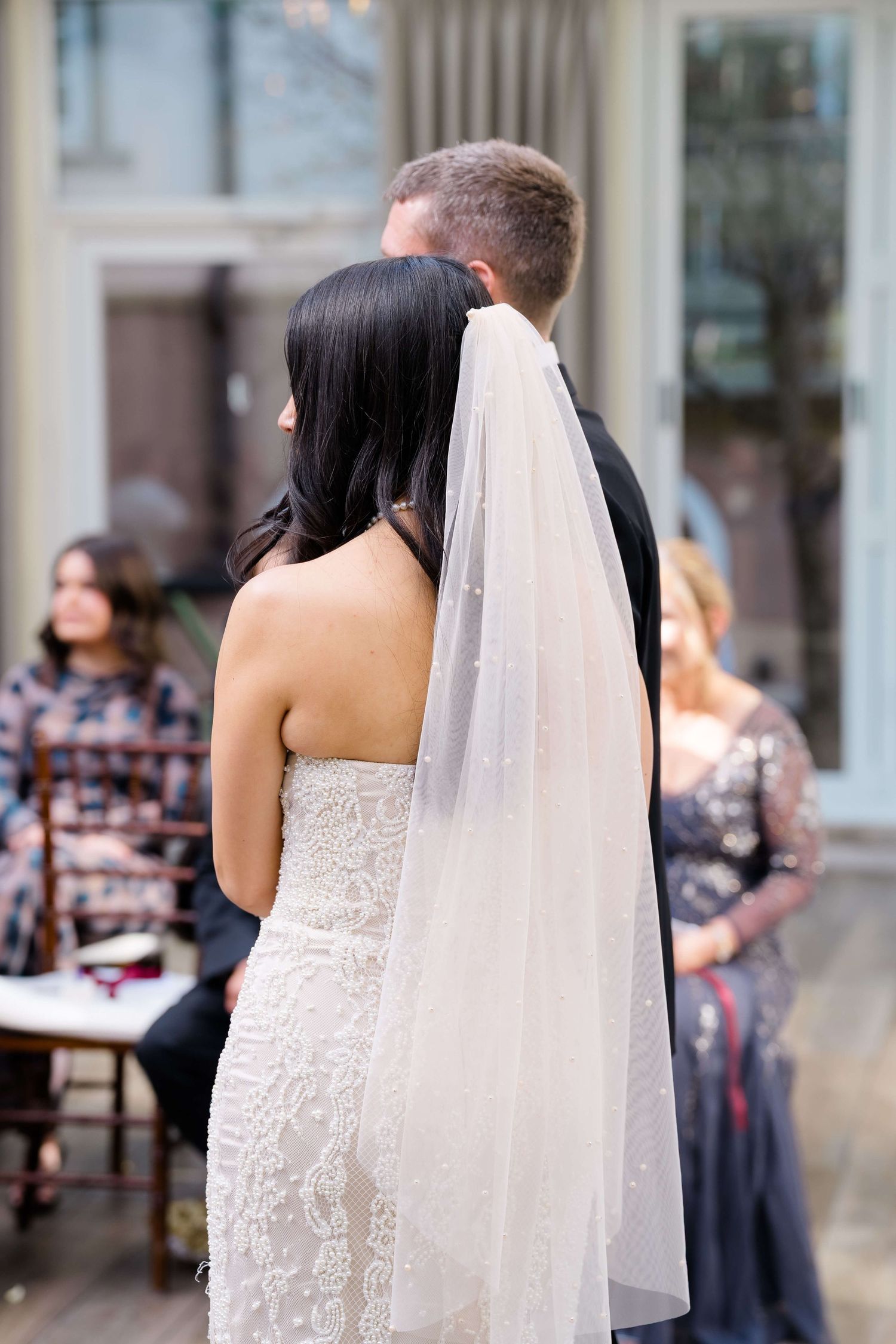 Bride in lace gown and veil stands beside groom during outdoor wedding ceremony with guests seated behind.