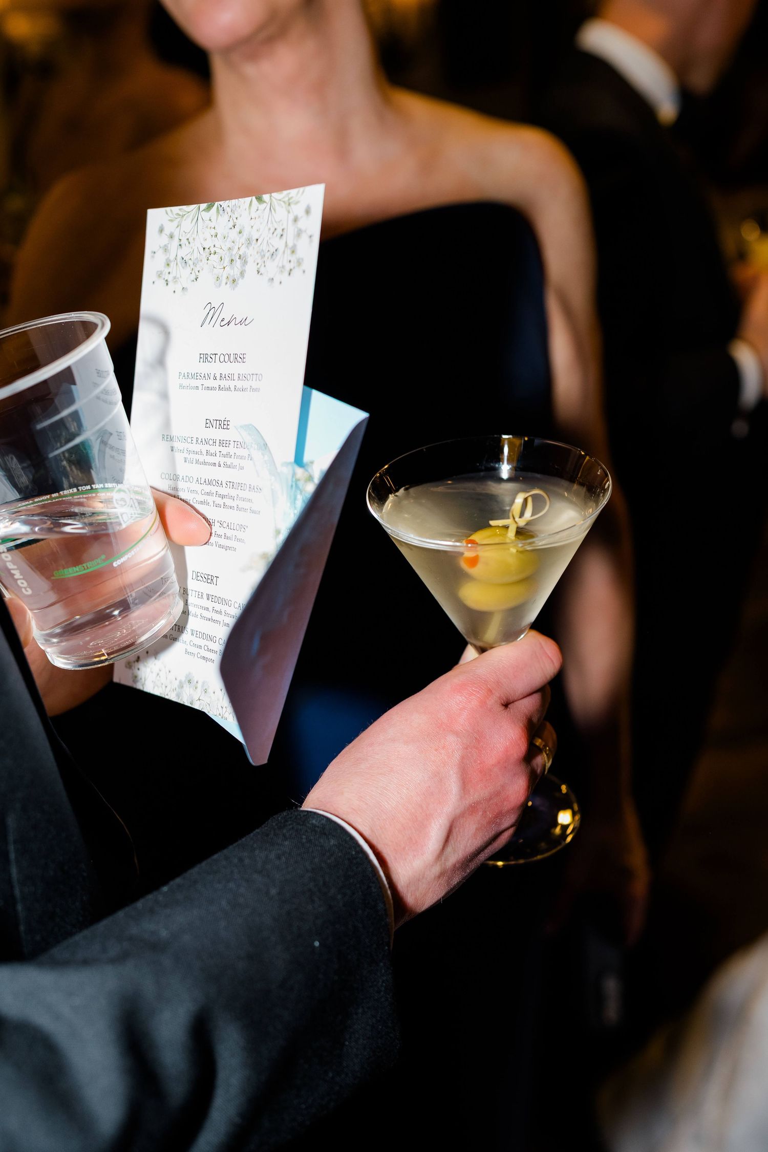 Person holding a dirty martini with olives and a wedding menu card at a formal event.
