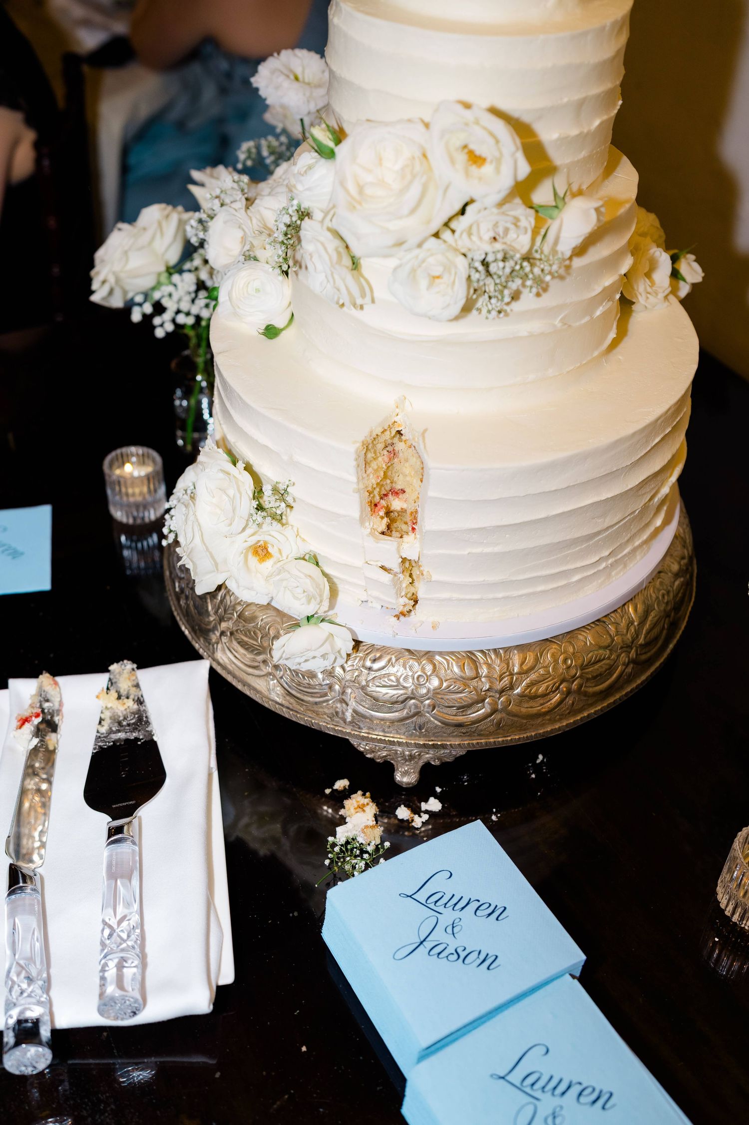 Elegant three-tier white wedding cake with roses on silver stand, beside cake cutting set and Lauren & Jason programs.