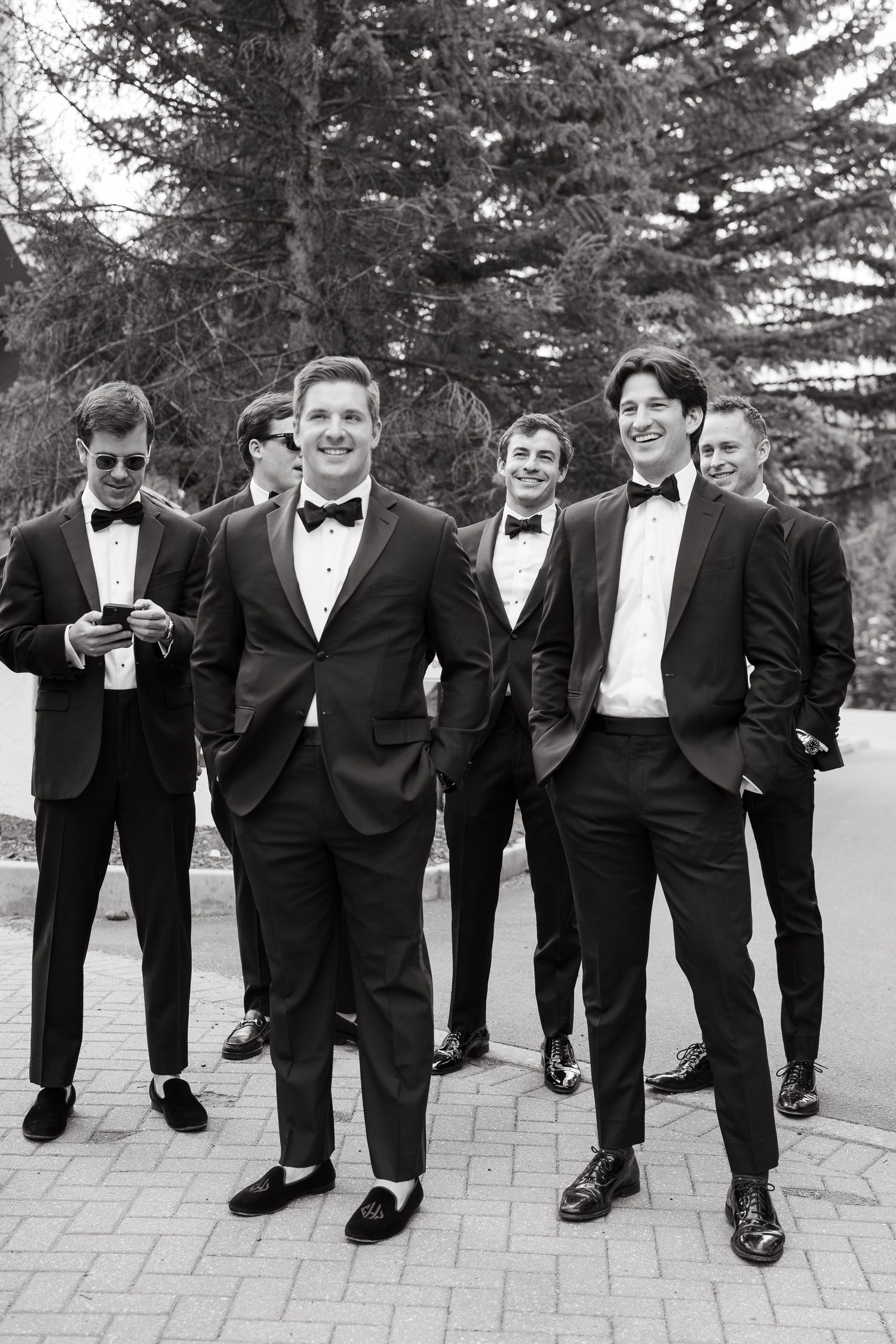 Black and white photo of a groom and groomsmen in tuxedos posing outdoors with trees in the background.