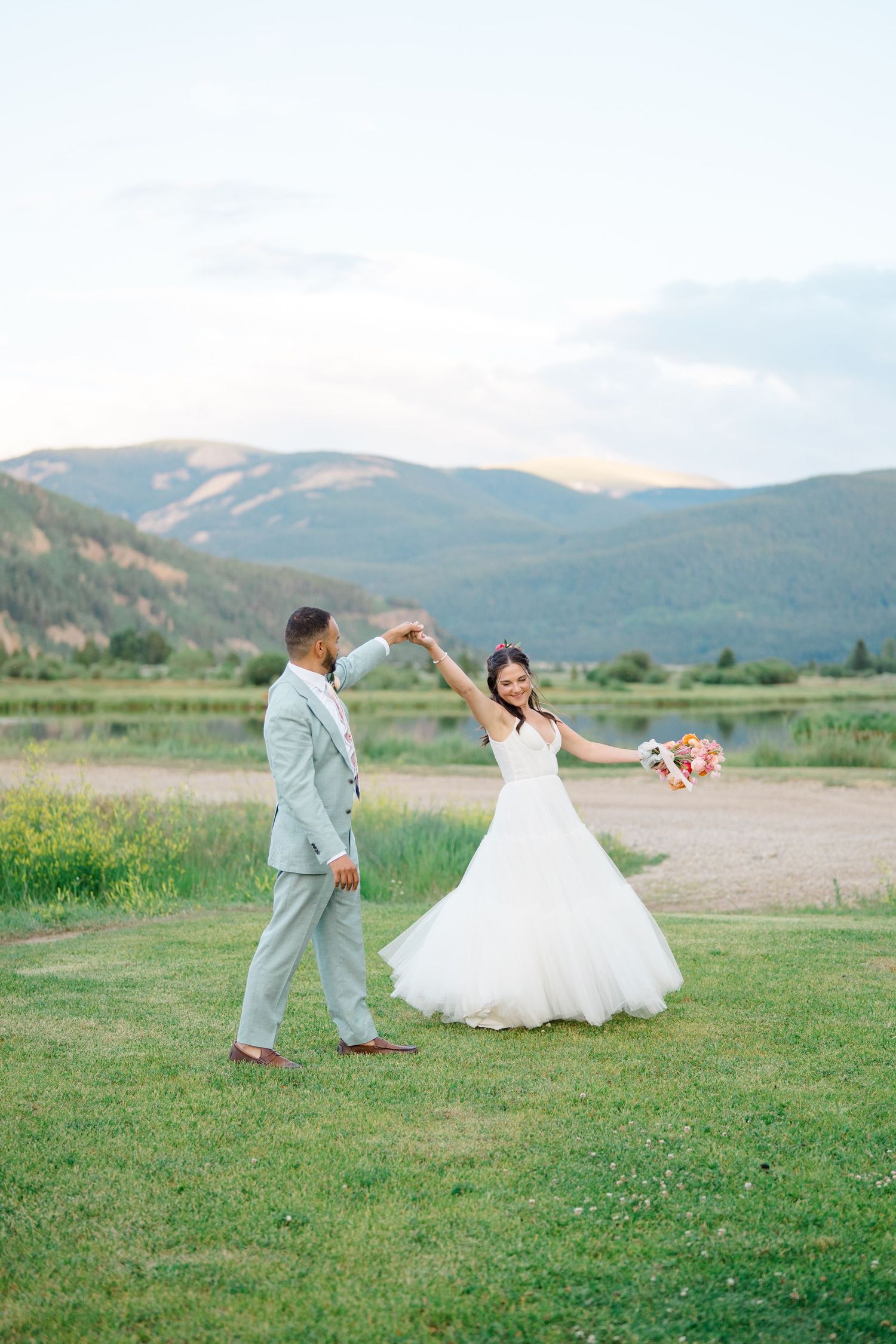 A couple dances playfully in a grassy mountain field during their wedding photoshoot.
