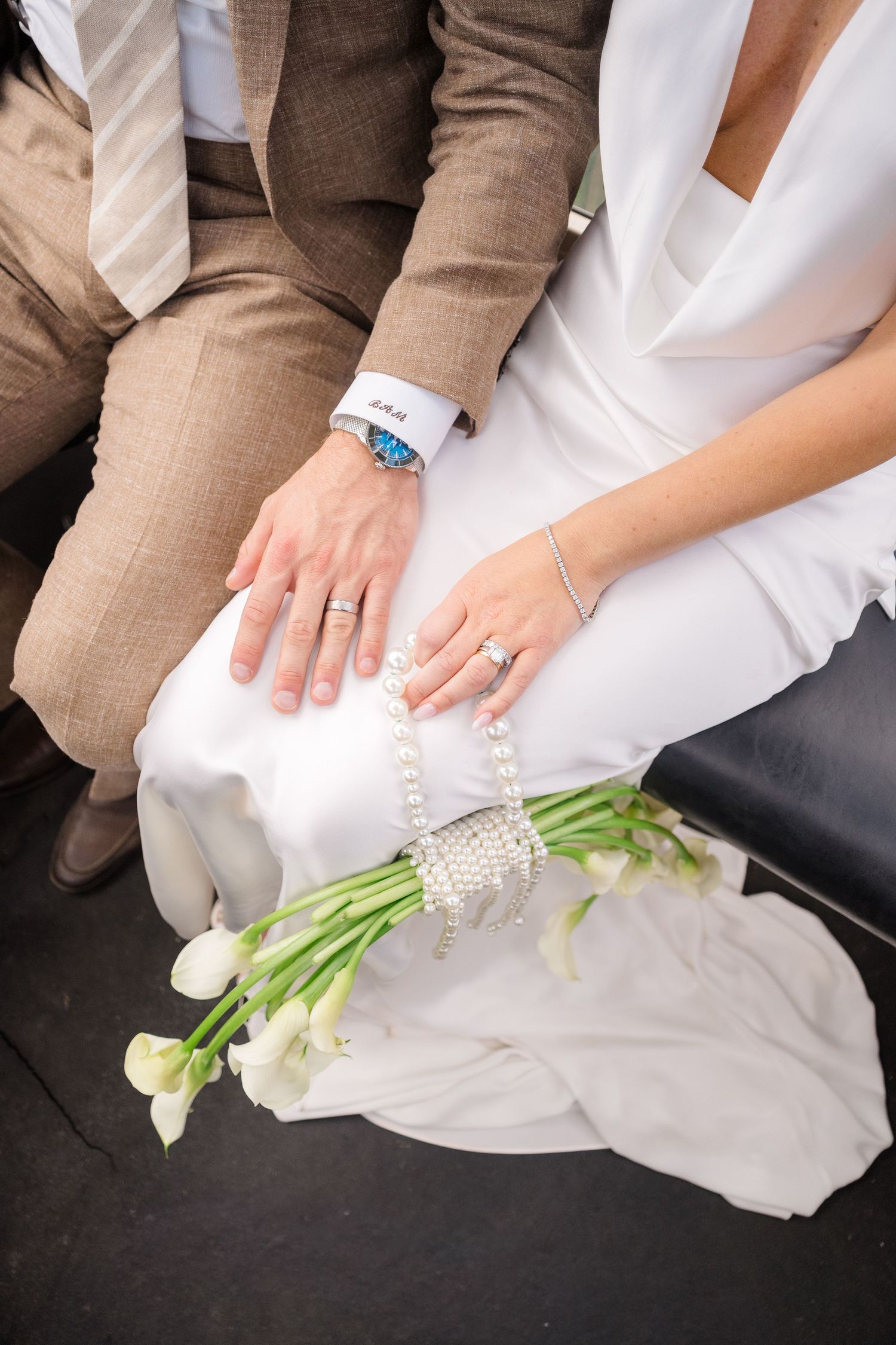 Bride and groom holding hands with wedding rings, pearl bouquet of white calla lilies on white dress.