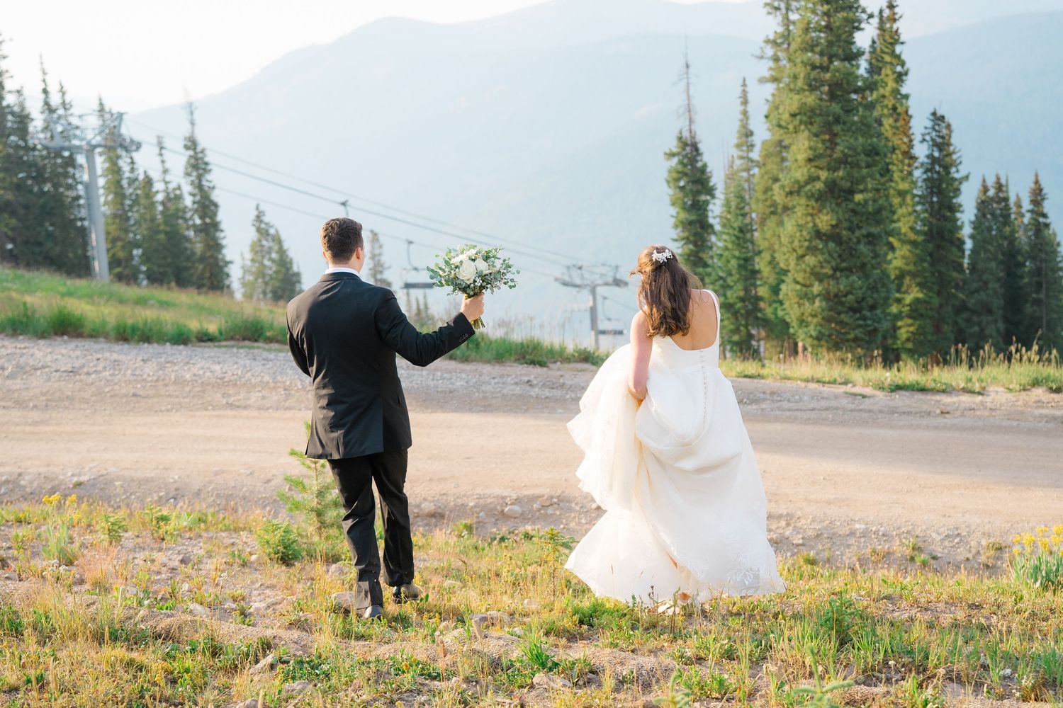Bride and groom walking hand in hand through mountain meadow, bride in white gown holding bouquet, evergreen trees backdrop.