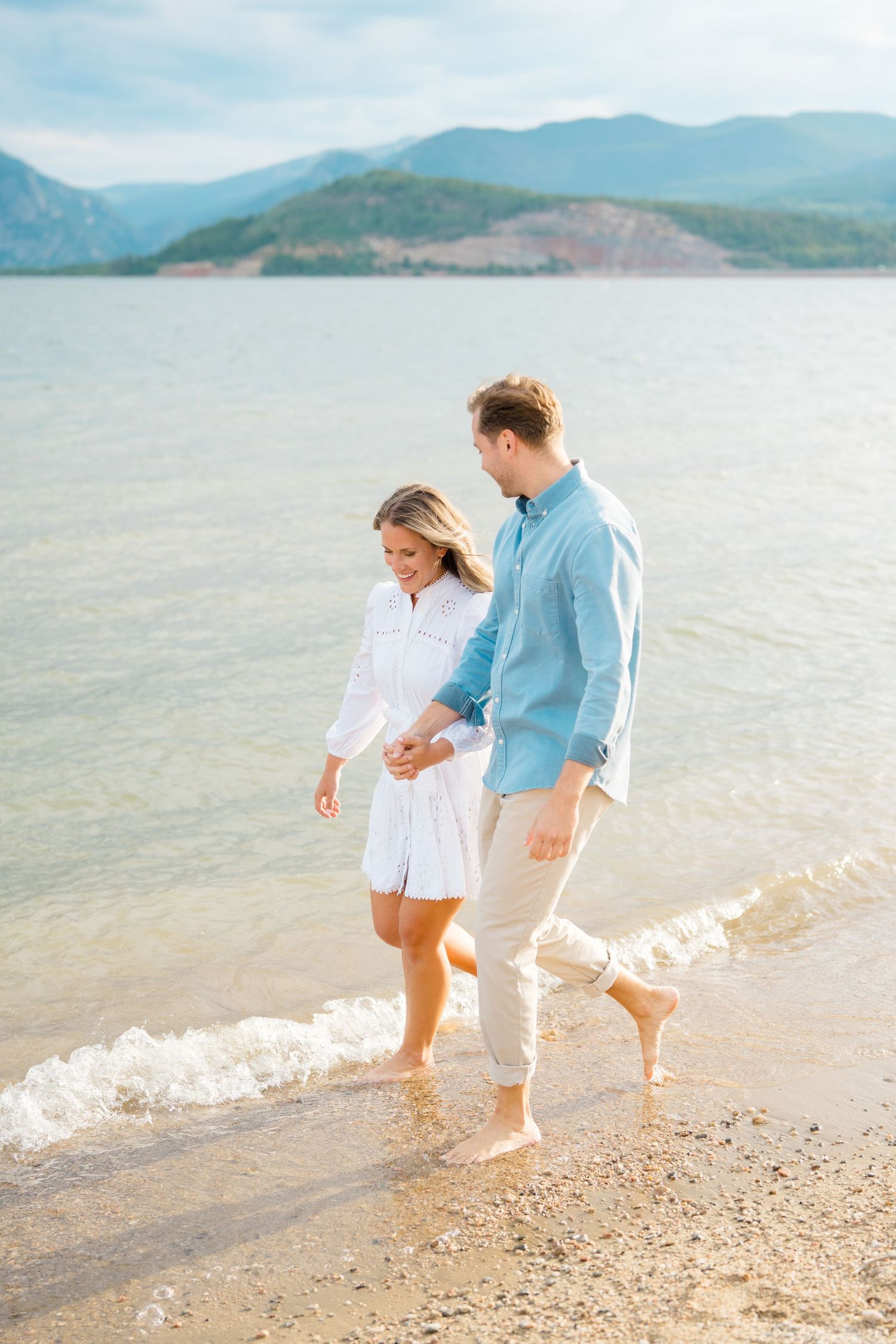 Couple walking together along a scenic beach shoreline with mountains in the background.