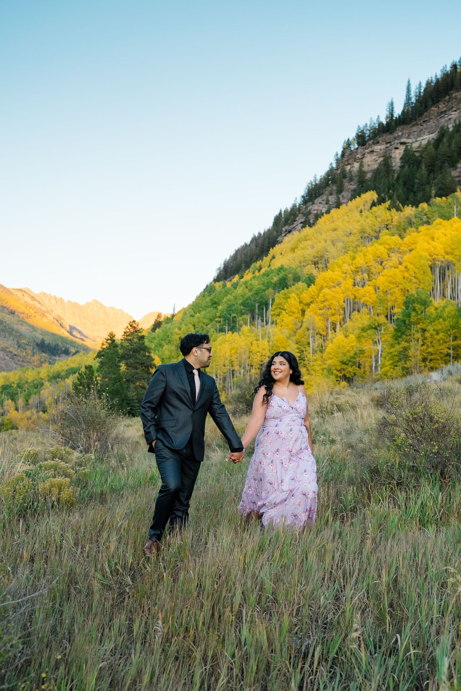 A couple walk hand in hand through a mountain meadow with golden aspen trees and cliffs in the background.