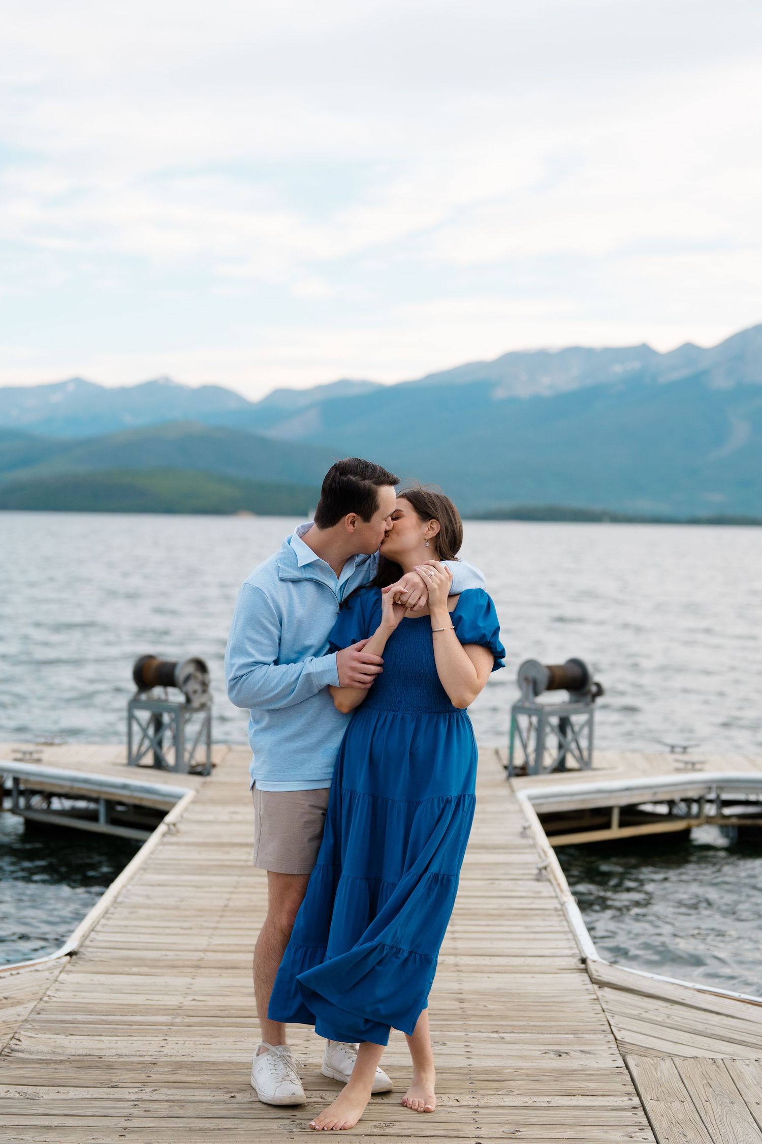 A couple shares a romantic moment on a wooden dock overlooking a mountain lake on a cloudy day.