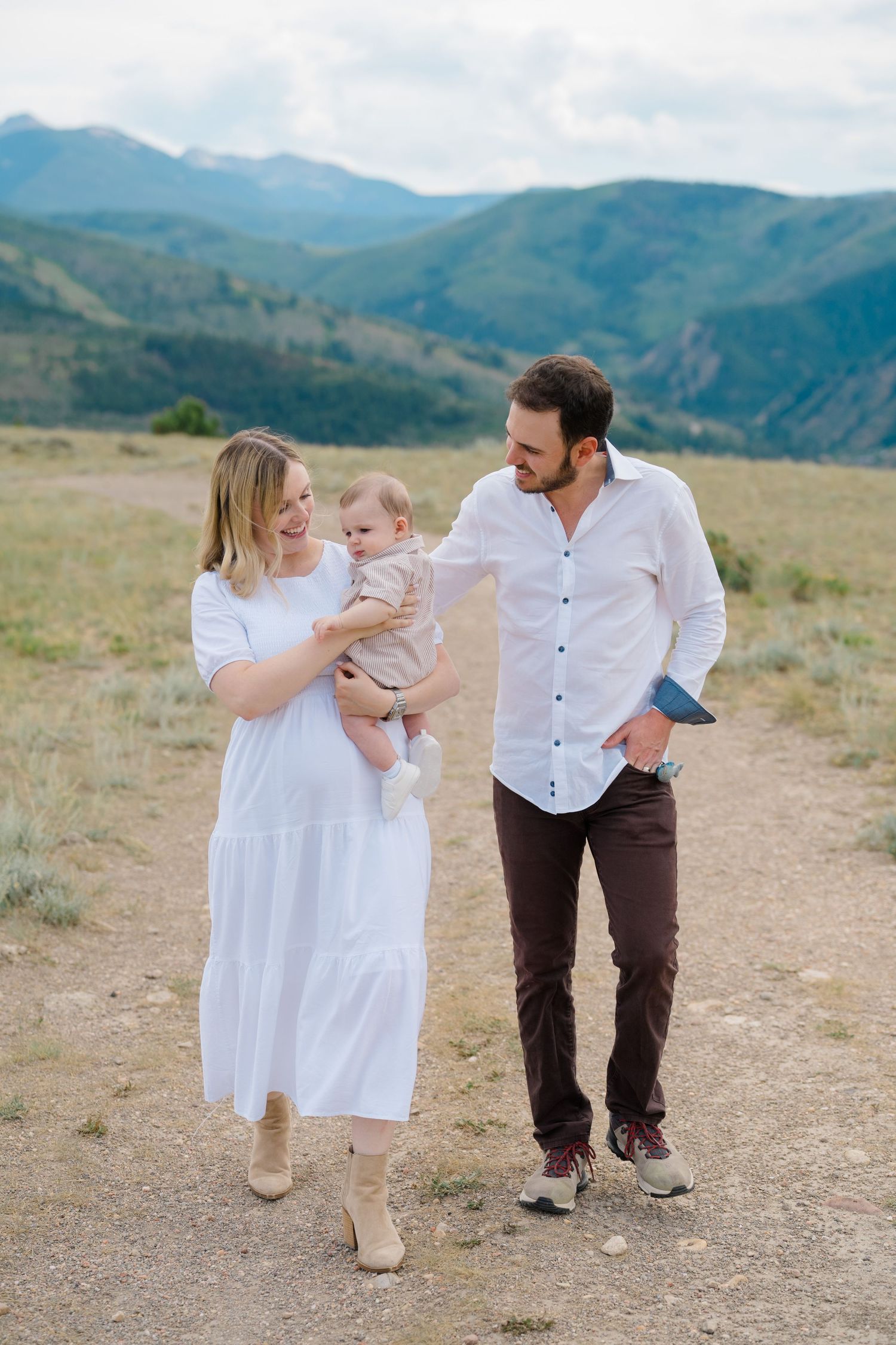 A couple walking with their baby along a mountain trail with scenic views in the background.