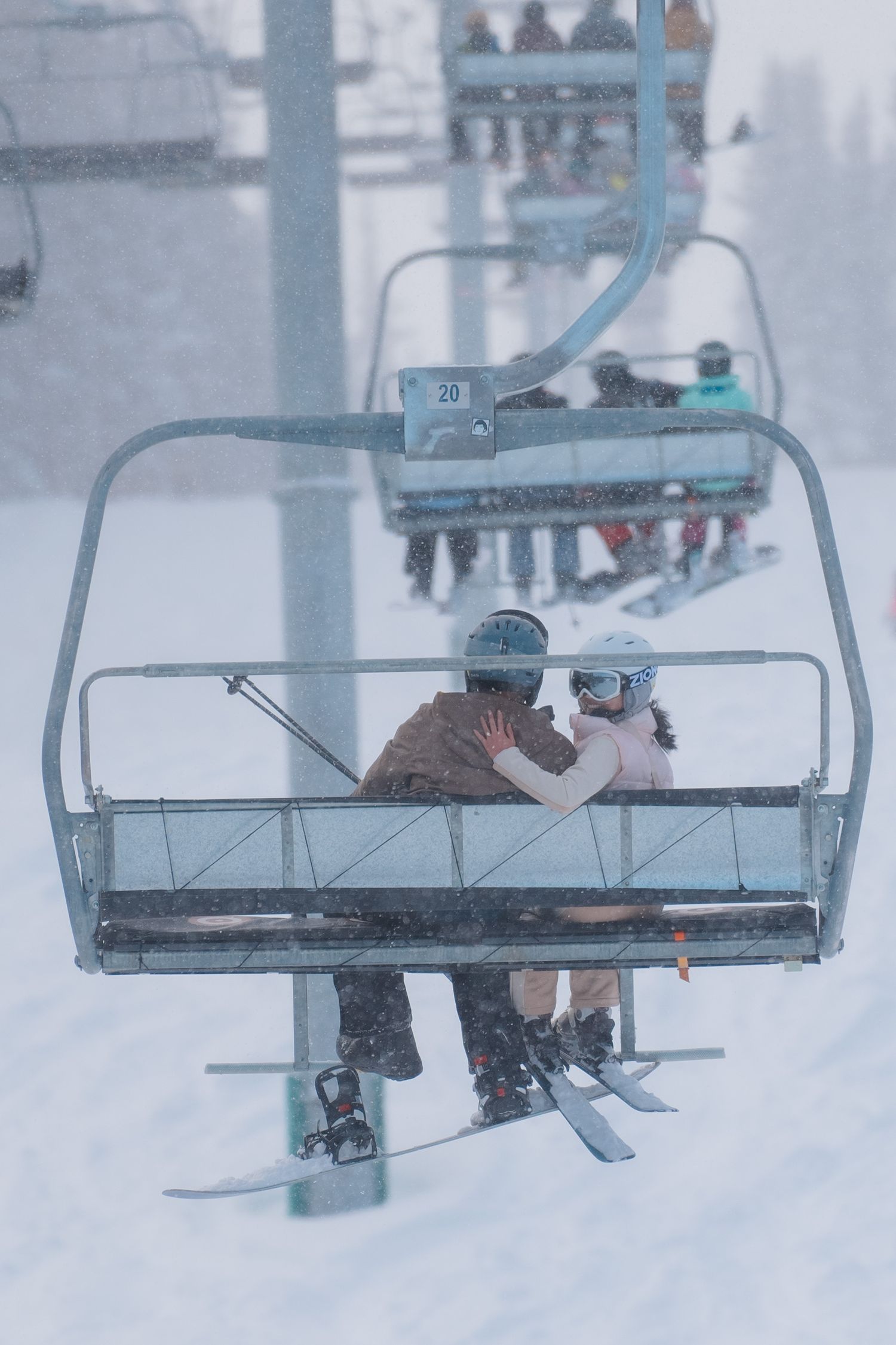 Skiers riding a chairlift up a snowy mountain slope during winter conditions with foggy weather.