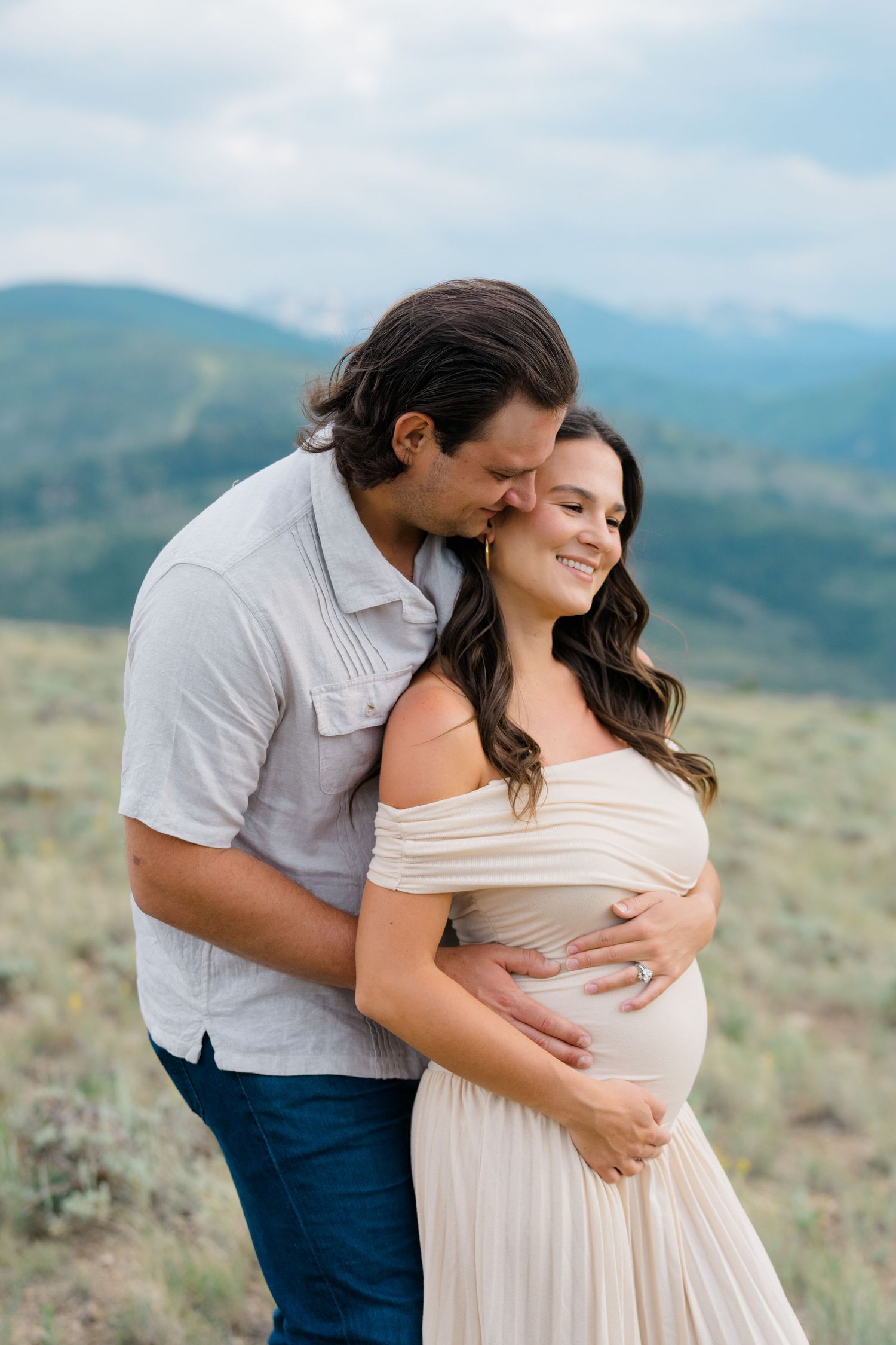 Couple embraces in a romantic maternity photoshoot against a scenic mountain landscape backdrop.