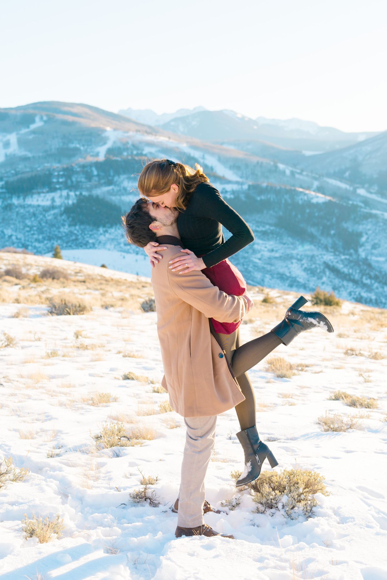 A couple embraces in a romantic pose against a snowy mountain landscape with blue winter skies in the background.