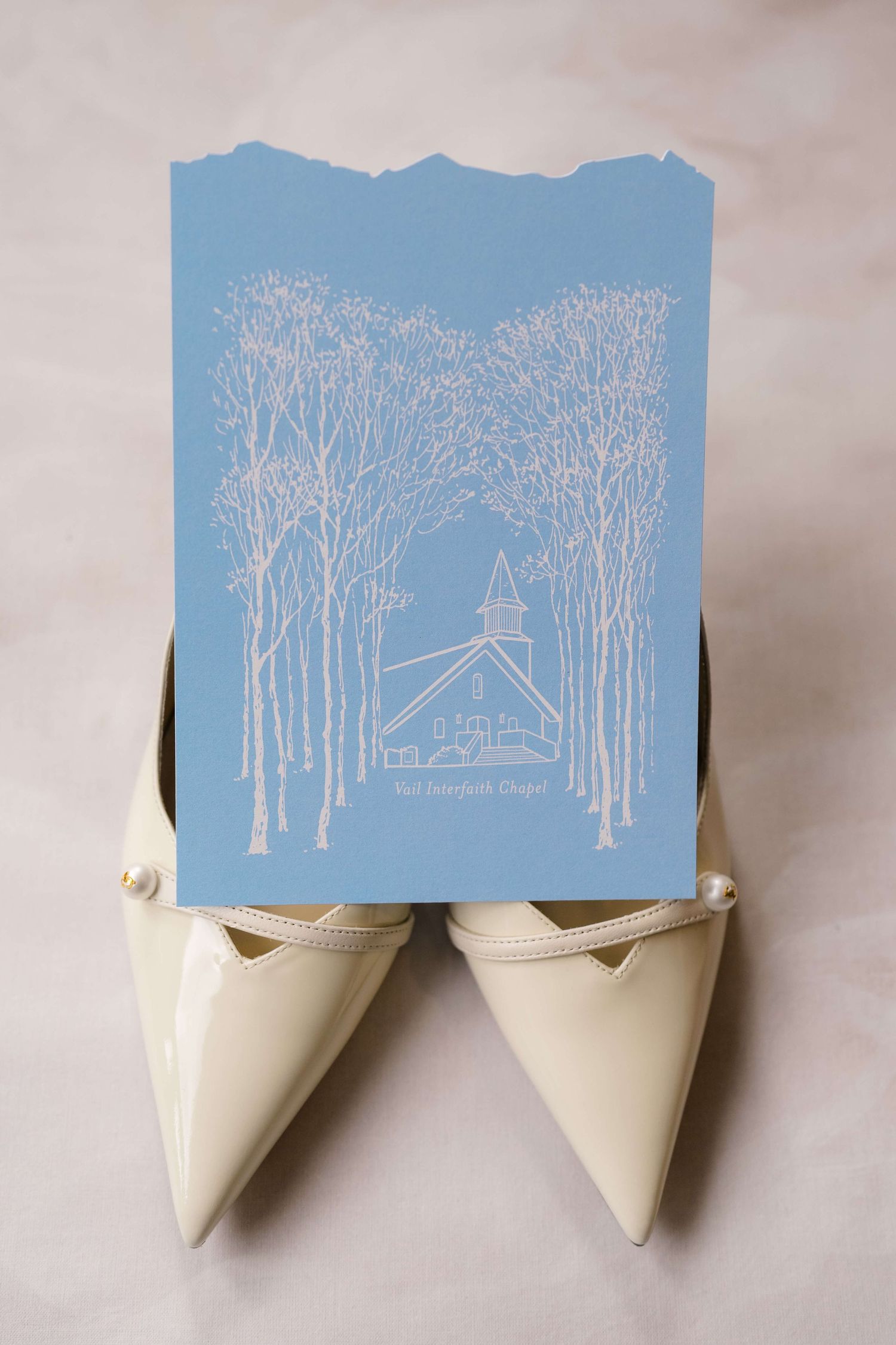 Blue wedding program card with church illustration leaning against ivory pointed-toe bridal heels on marble surface.