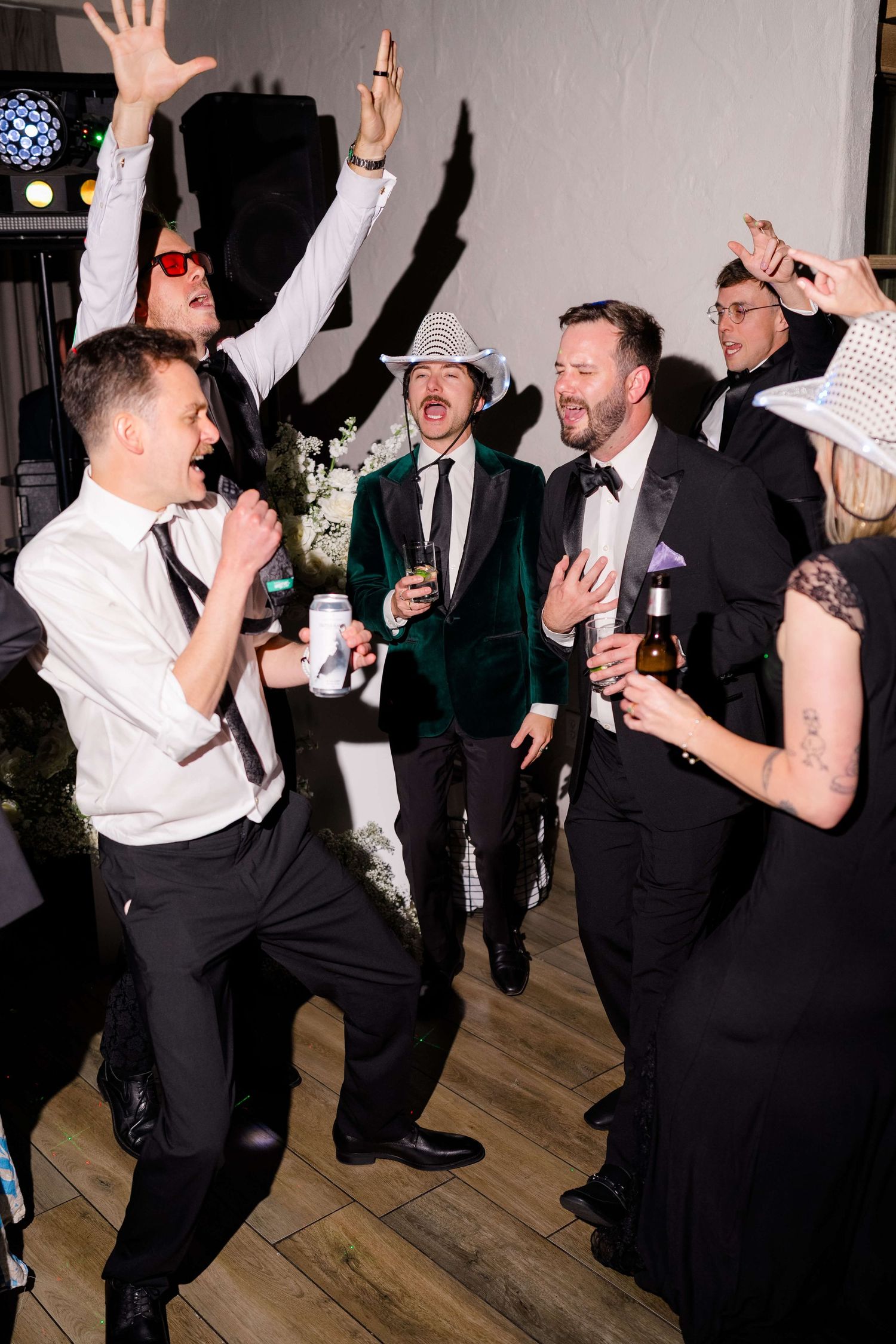 Guests dancing and celebrating at a lively wedding reception, wearing formal attire and festive hats.