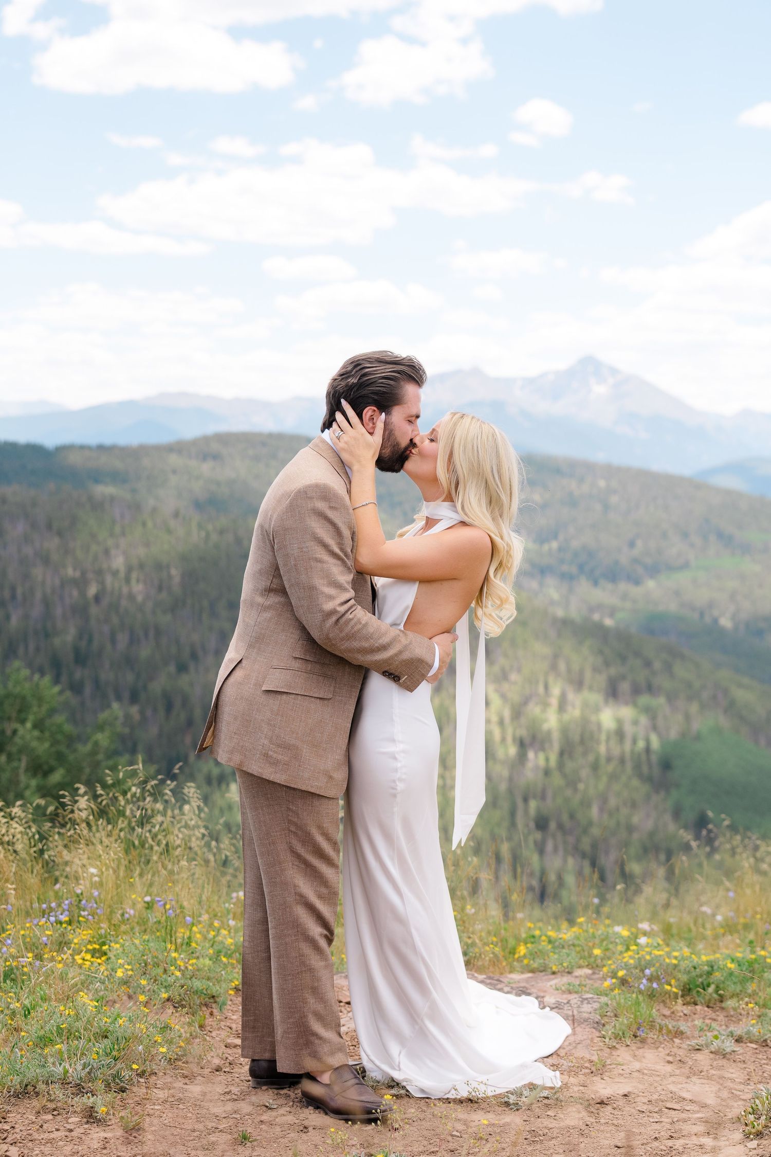 A bride and groom share a kiss on a mountain overlook, surrounded by wildflowers and scenic Colorado peaks.