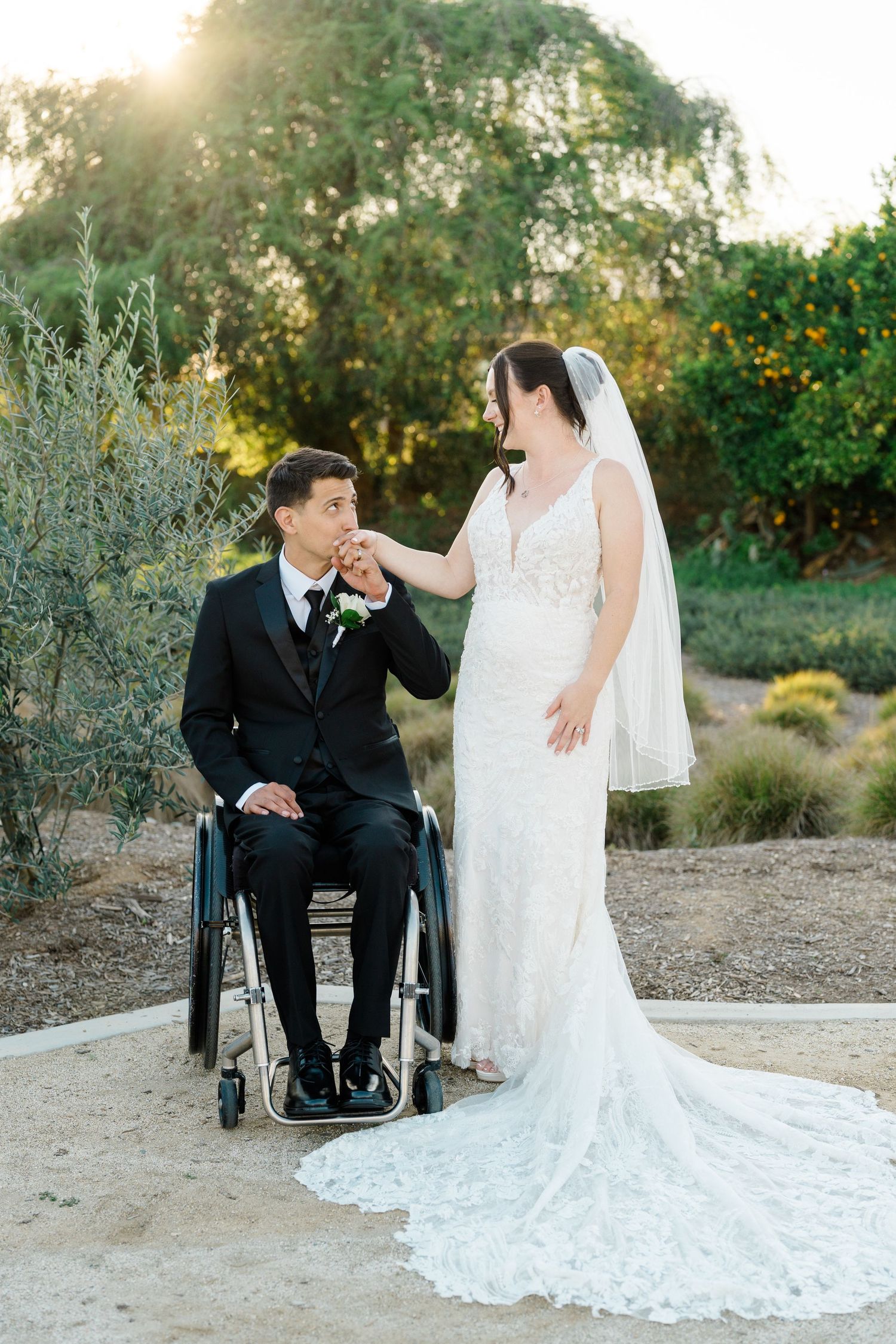 Bride in lace gown holds groom's hand as he sits in wheelchair, sharing a romantic moment outdoors at golden hour.