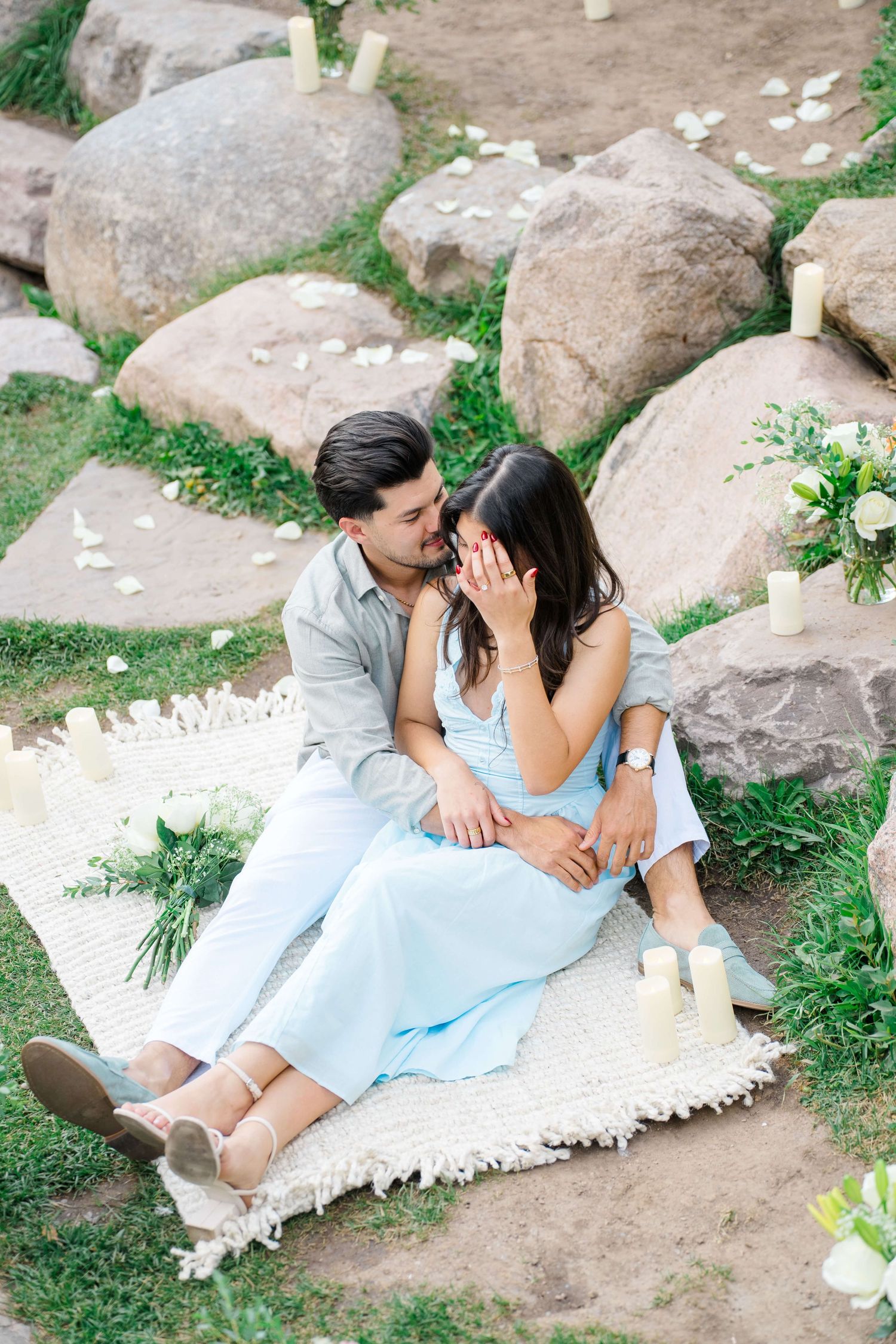 A romantic couple sits on a blanket among rocks and greenery in soft pastel outfits during an outdoor proposal in vail village.