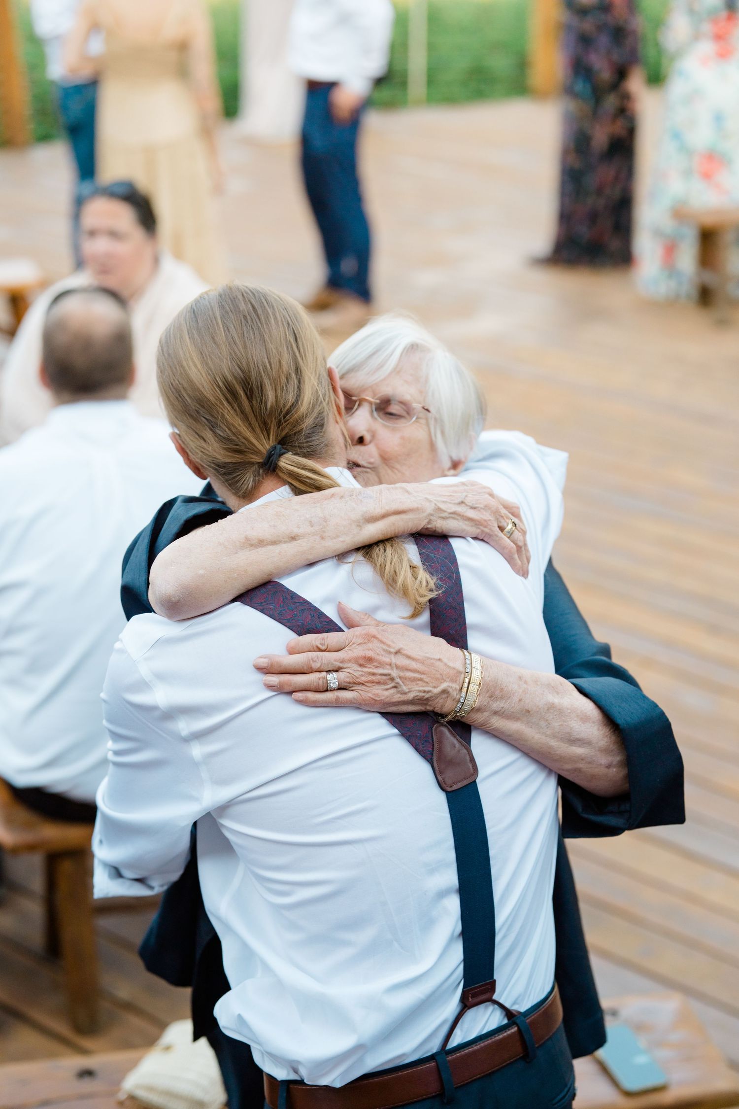 Wedding guests share emotional embraces on a wooden deck during celebration.